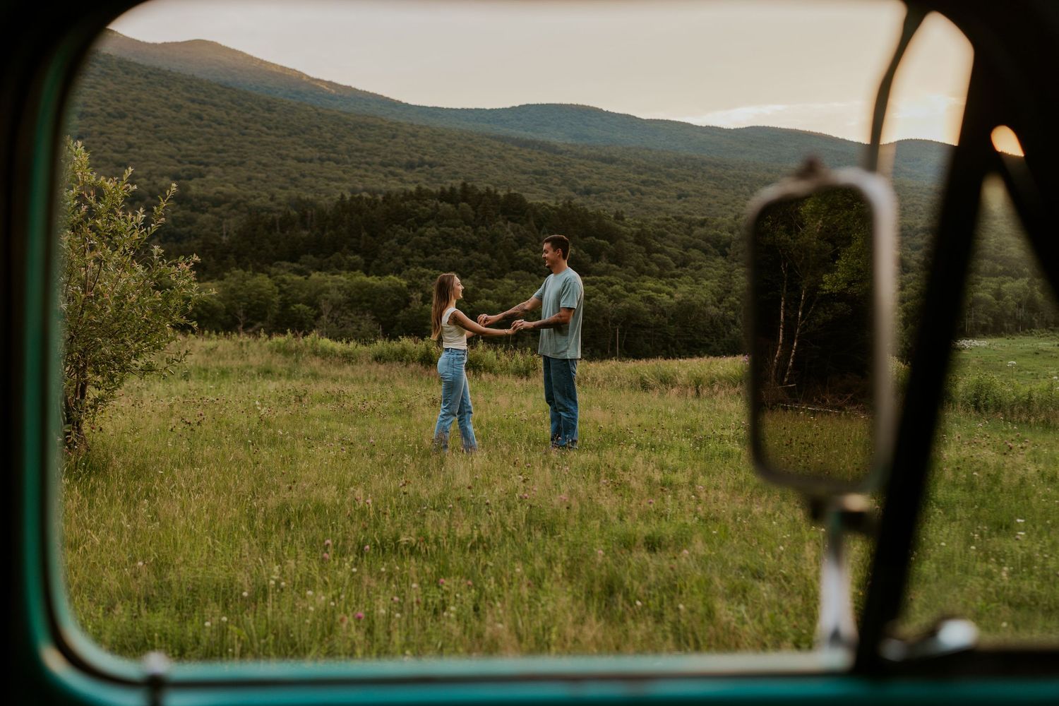 Couple dancing in a lush green field with mountain views, framed through a vintage truck window.