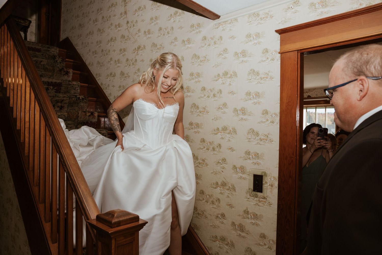 Bride in strapless white gown descending wooden staircase with wallpapered walls on her wedding day.