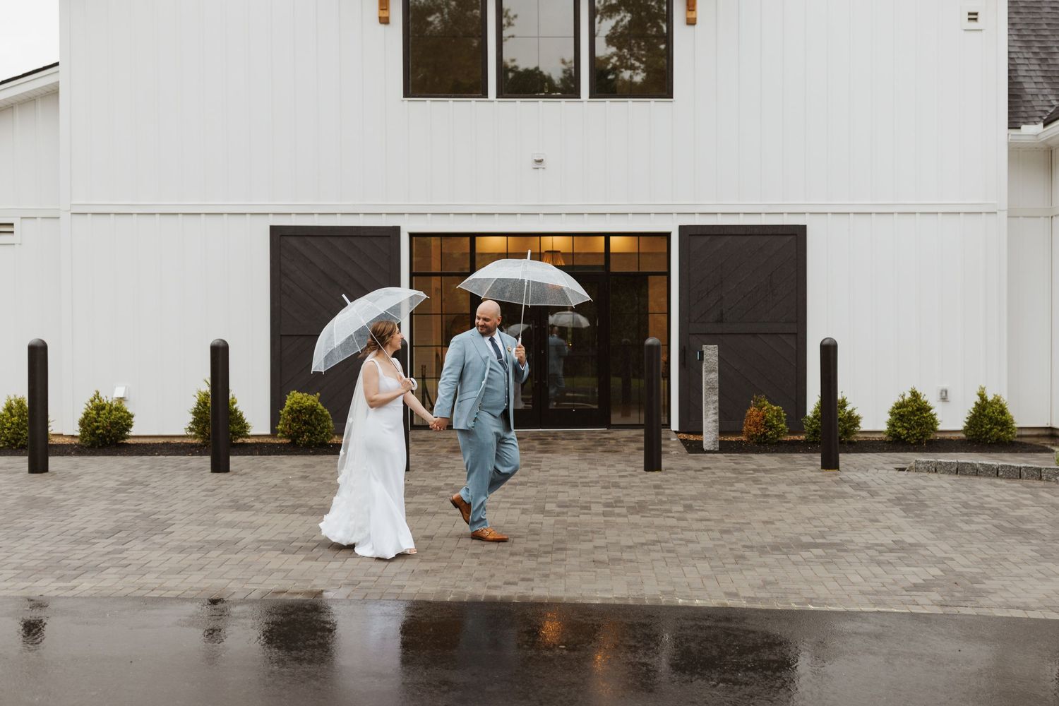 Bride and groom walking hand in hand with umbrellas outside a white barn in rain.