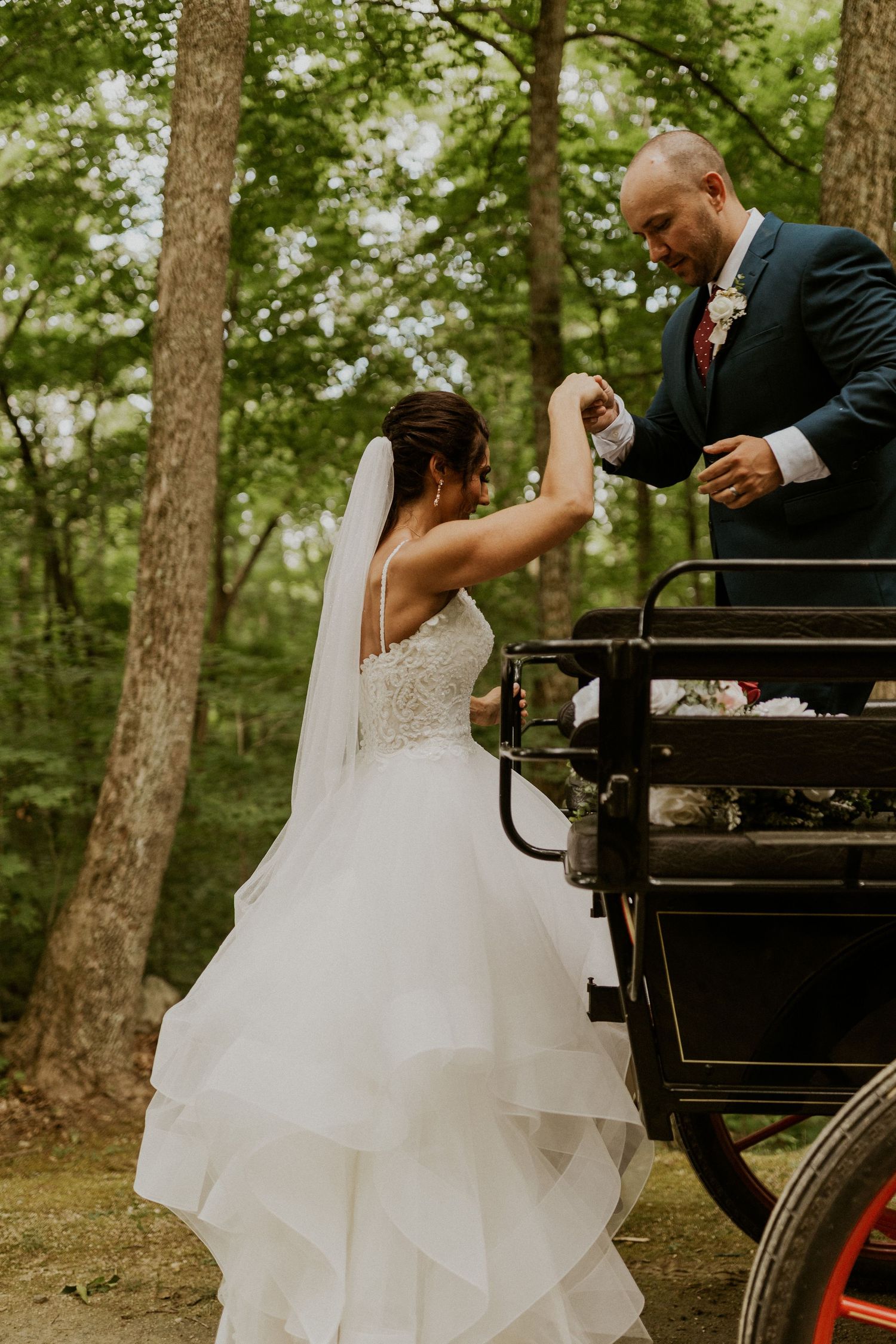Elegant white wedding dress flowing as couple boards a vintage horse-drawn carriage in the woods.