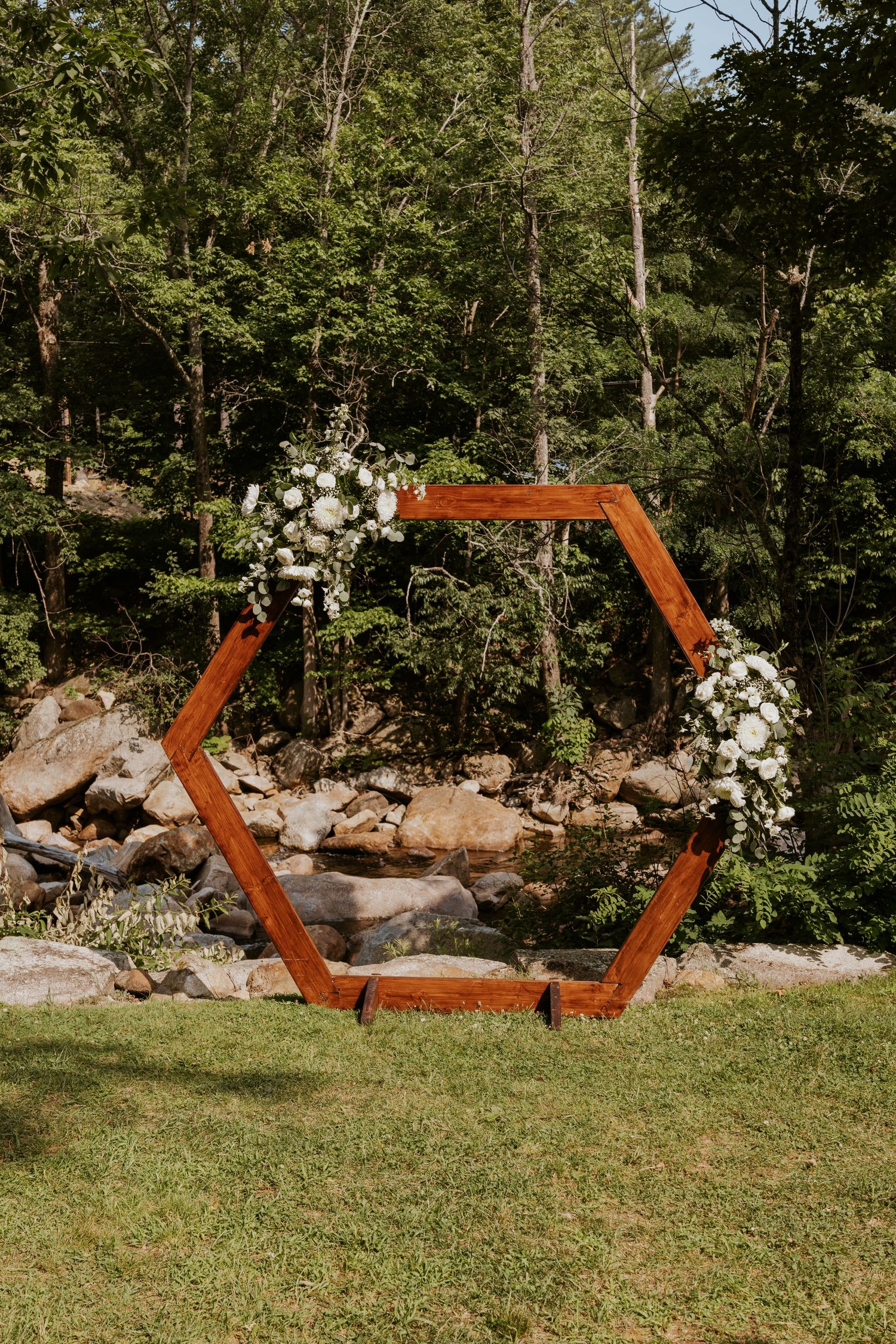 A rustic hexagonal wooden wedding arch adorned with white florals stands outdoors near a rocky stream.