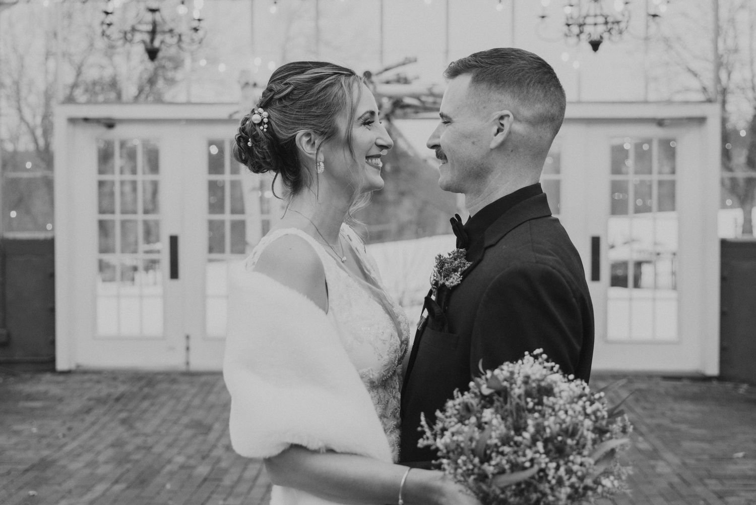 Black and white photo of smiling bride and groom embracing outside elegant white wedding venue doors.