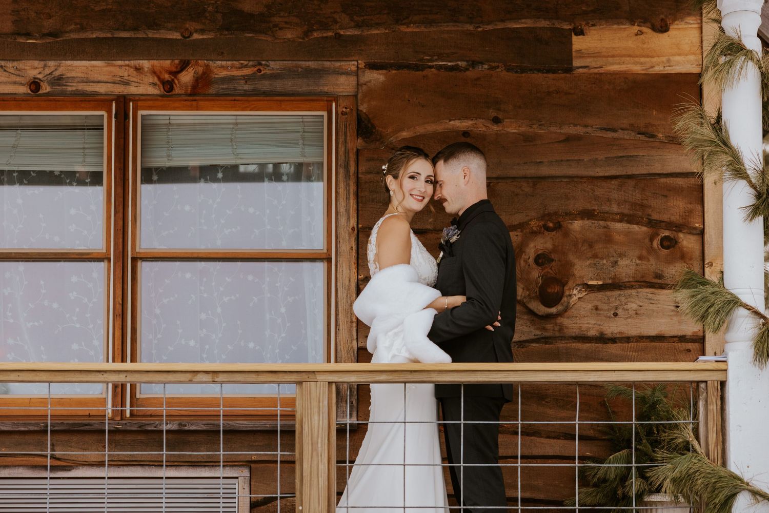 Bride and groom embrace on rustic log cabin balcony in winter, bride wearing white gown with fur wrap.
