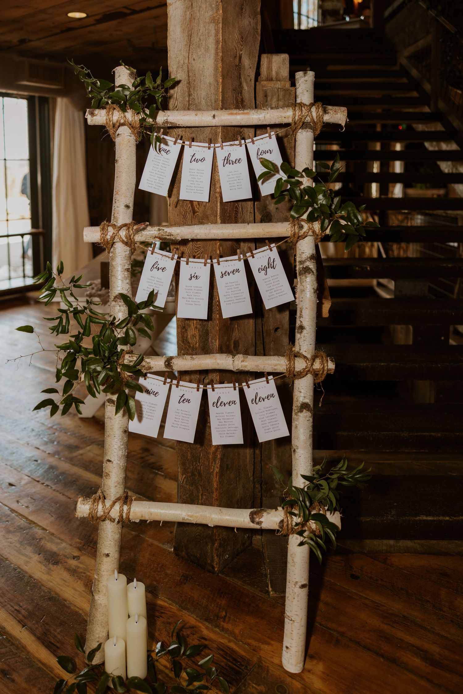 Rustic wedding seating chart display with white cards hung on birch poles decorated with greenery and candles.