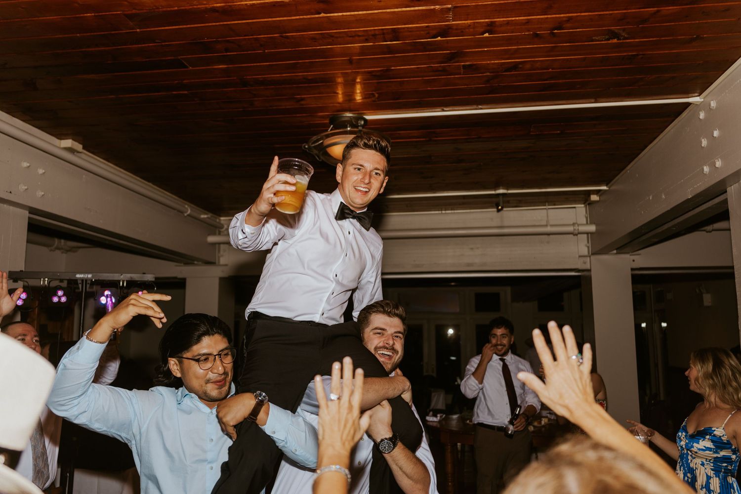 Wedding guest lifted on shoulders holding drink during lively reception party.