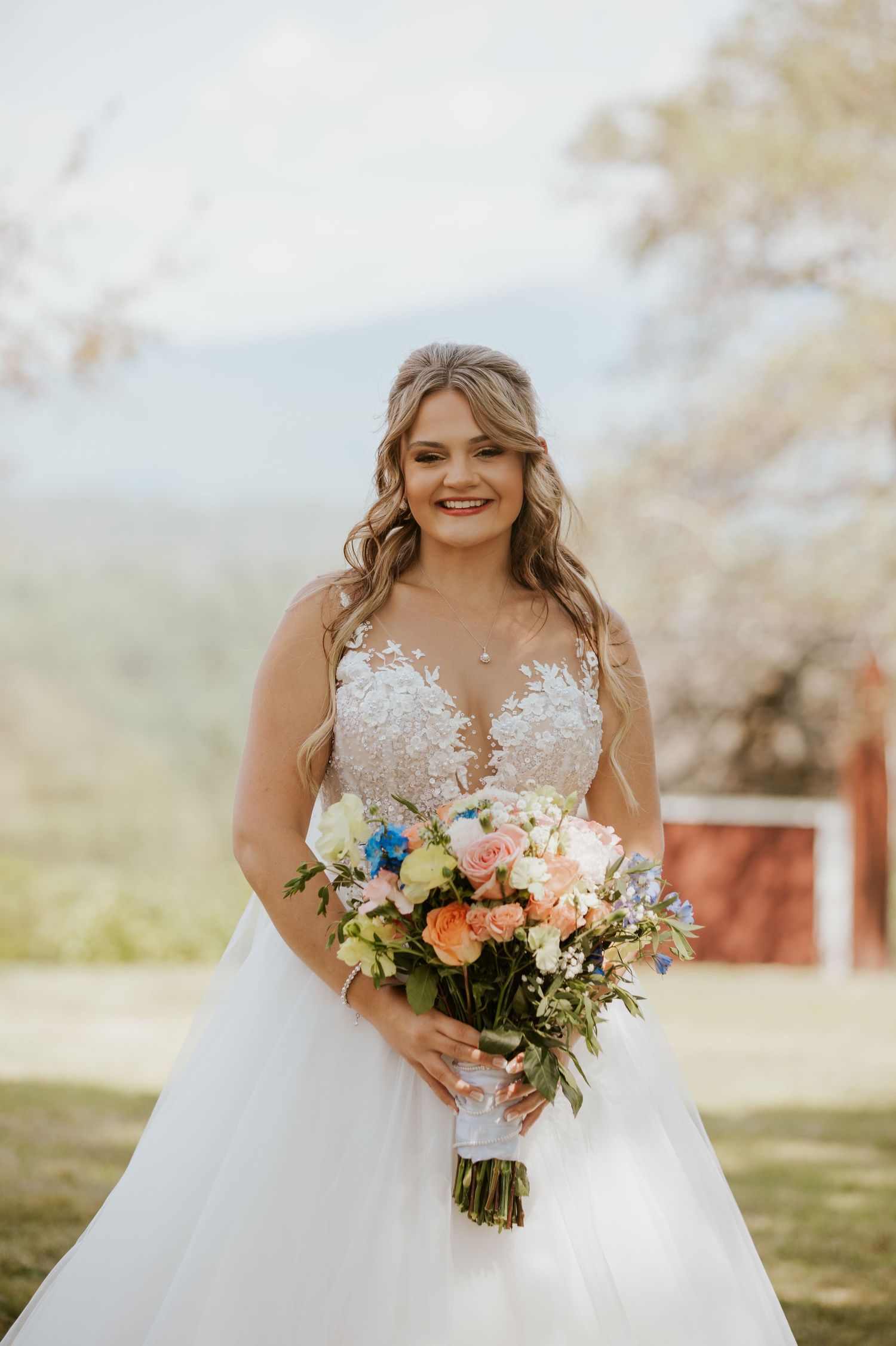 Smiling bride in lace ball gown holds colorful bouquet with mountain backdrop on wedding day.