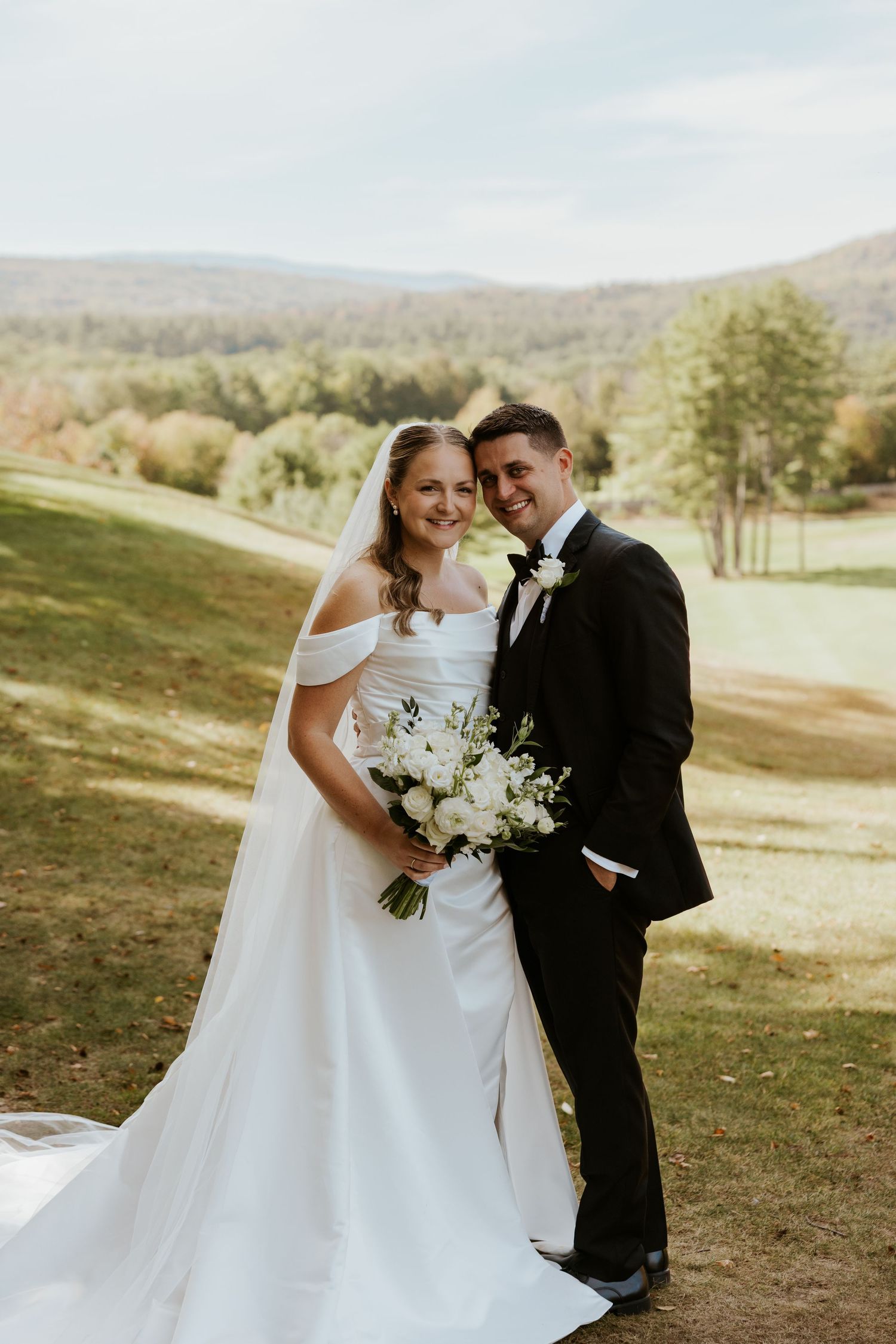 Smiling bride and groom pose together on a lush golf course with rolling mountains in the background.