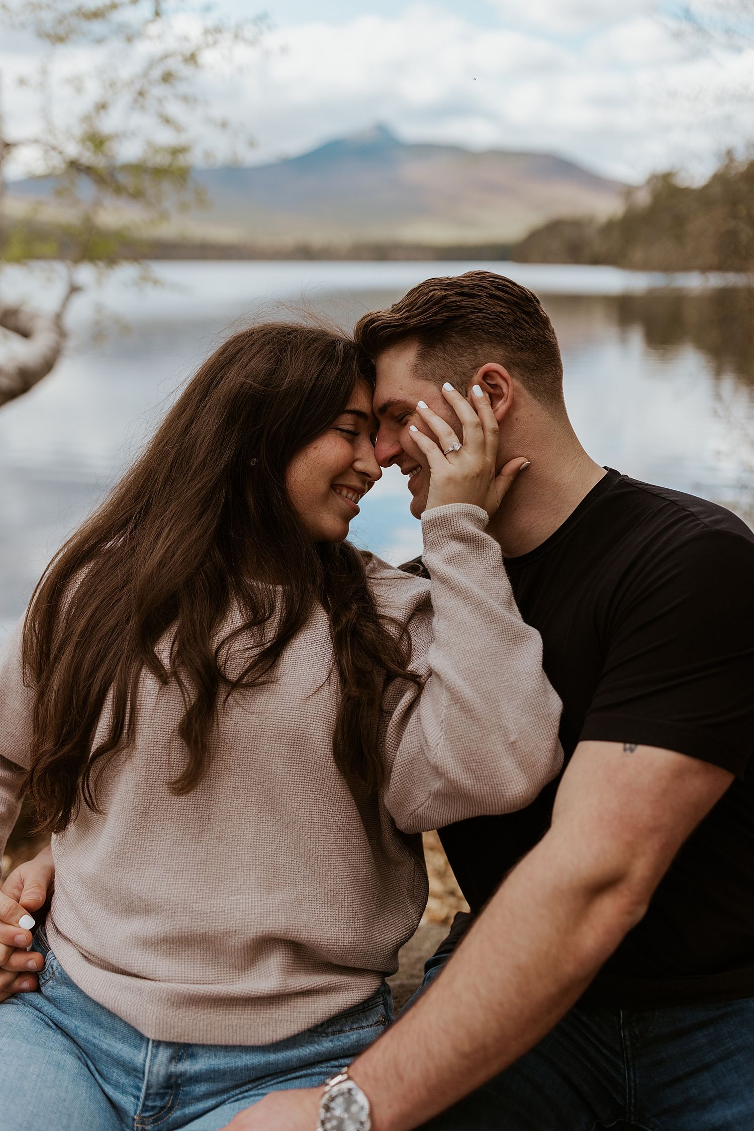 Romantic couple touching foreheads and smiling by a serene lake with mountains in the background.