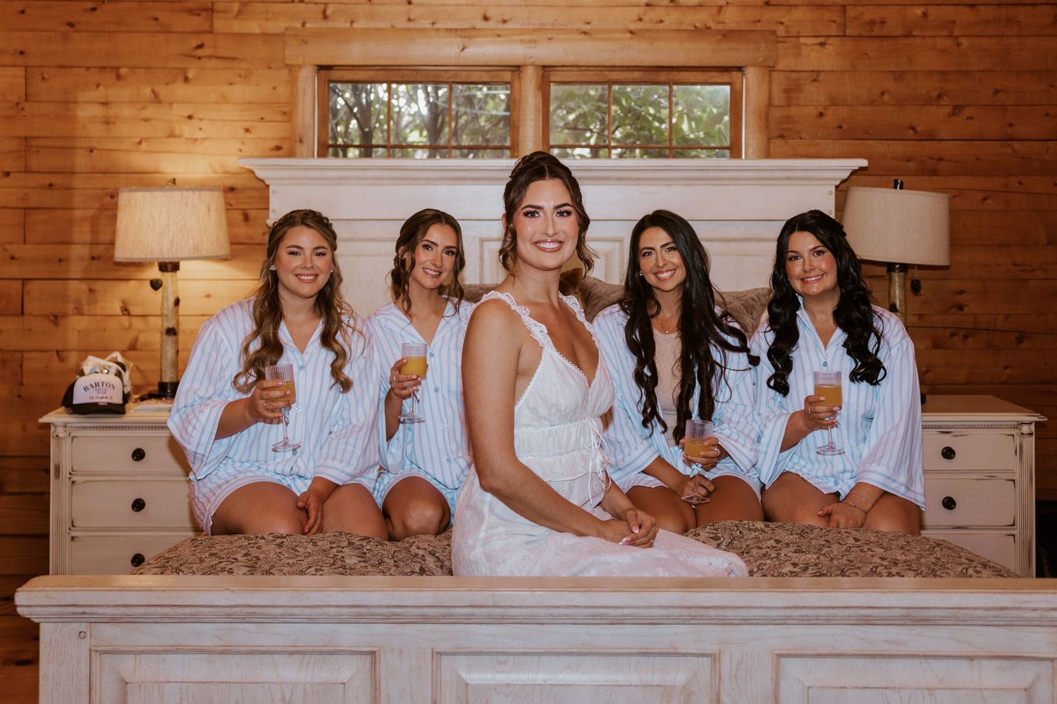 Bride and four bridesmaids in robes sitting on a white bed holding champagne.