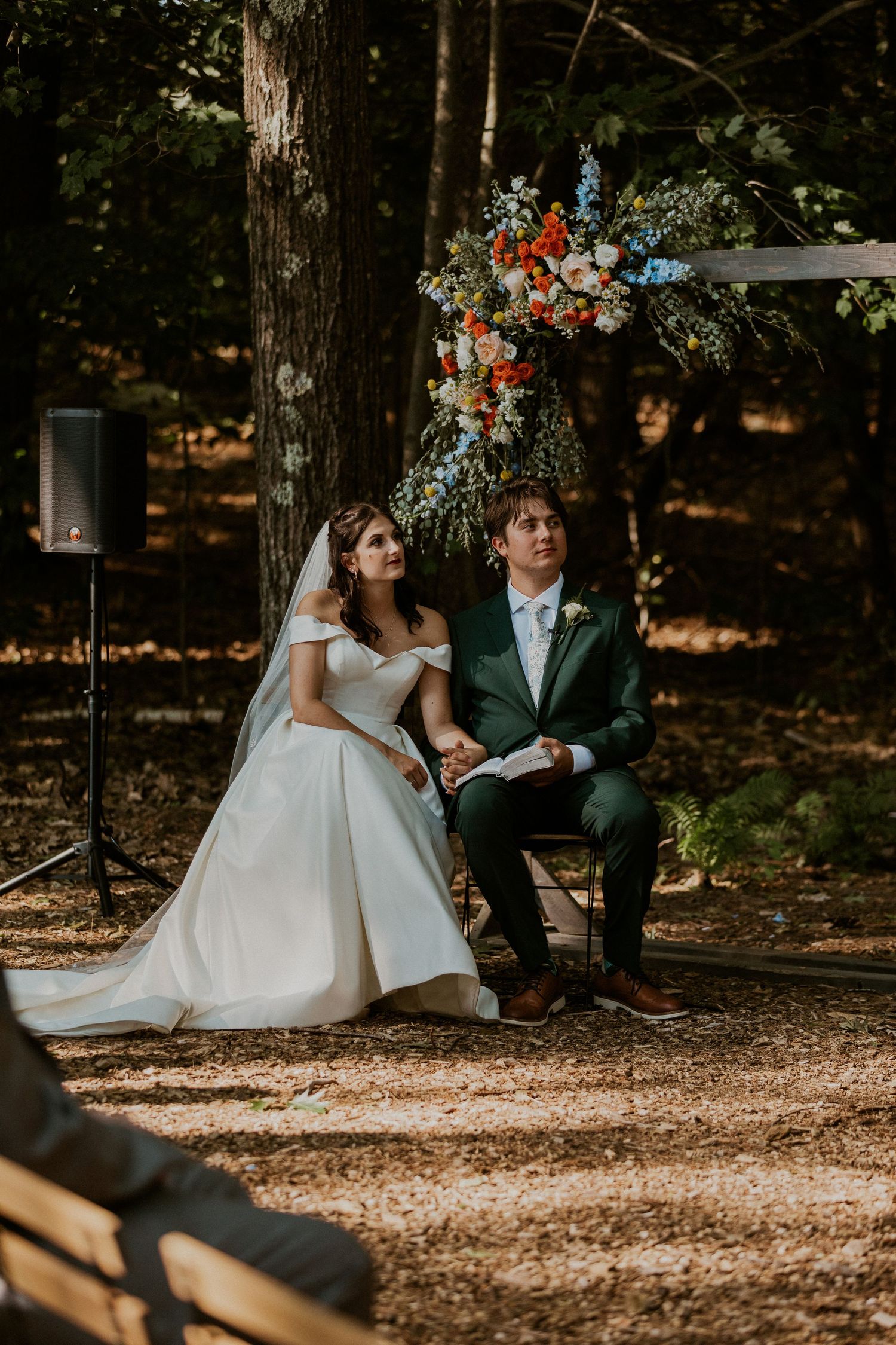 Couple seated during their outdoor woodland wedding ceremony with floral decorations.