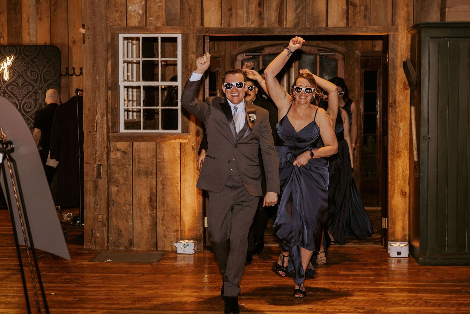 Groomsman and bridesmaid make a celebratory entrance at a rustic barn wedding reception with arms raised.