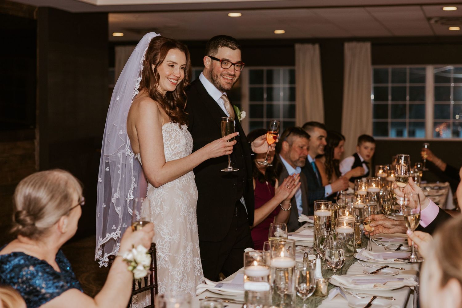 A wedding reception dinner with candlelit tables and guests raising glasses for a toast.