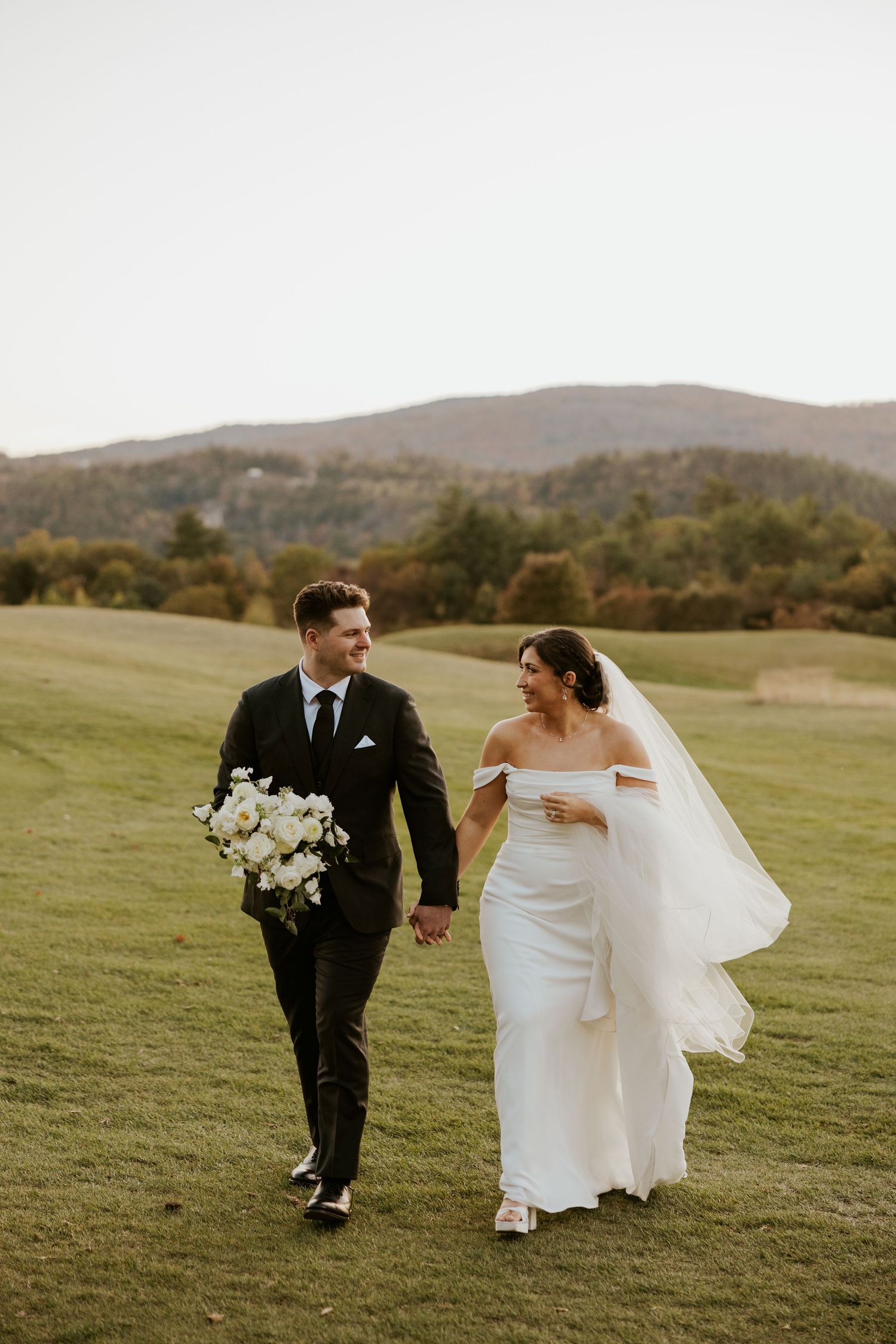 Bride in off-shoulder gown and groom in black suit walk hand-in-hand on a golf course with mountain views.