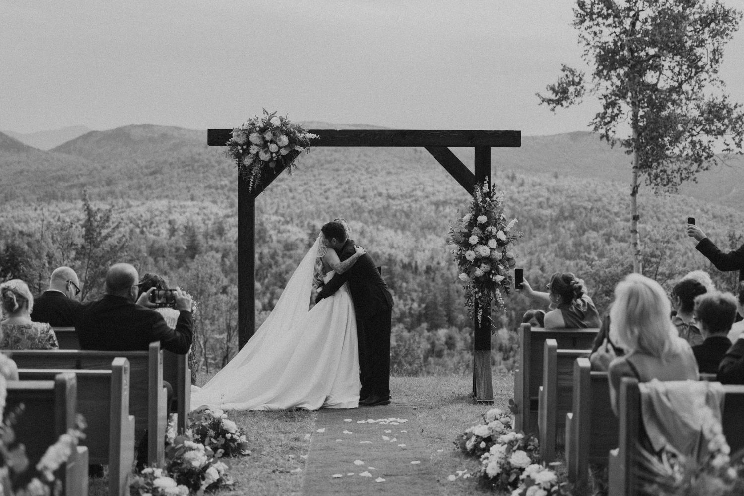 Black and white outdoor wedding ceremony with couple kissing under floral arch, mountain landscape backdrop, guests seated on wooden pews.