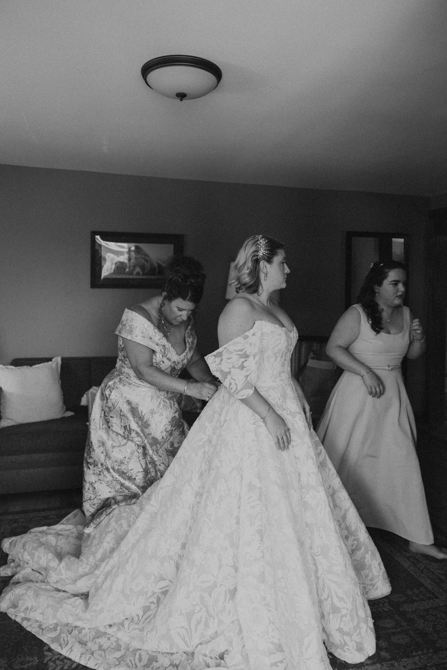 Black and white photo of bride being helped into her lace ball gown wedding dress.