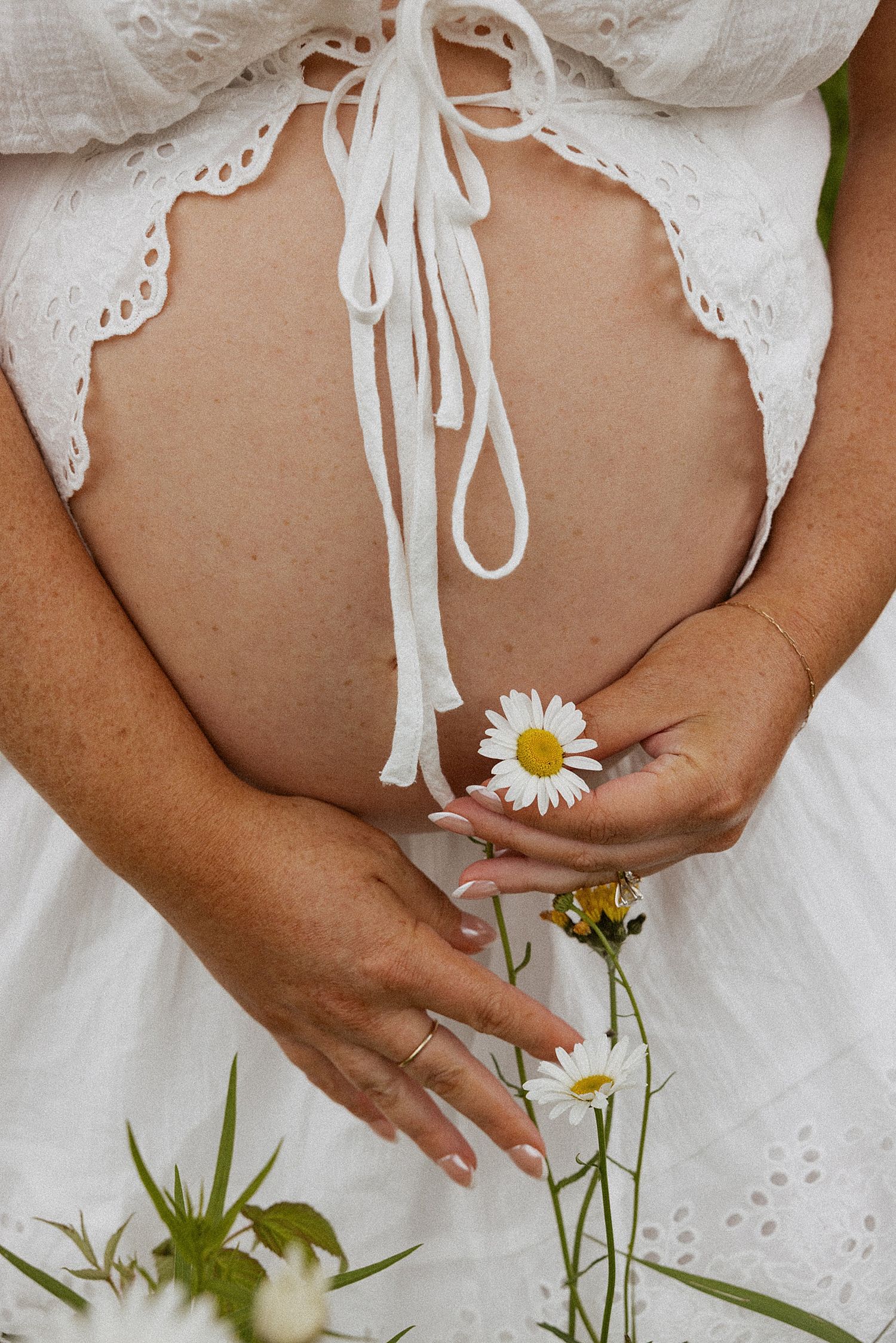 Pregnant woman in white lace dress holding daisy flowers against her baby bump outdoors in New Hampshire