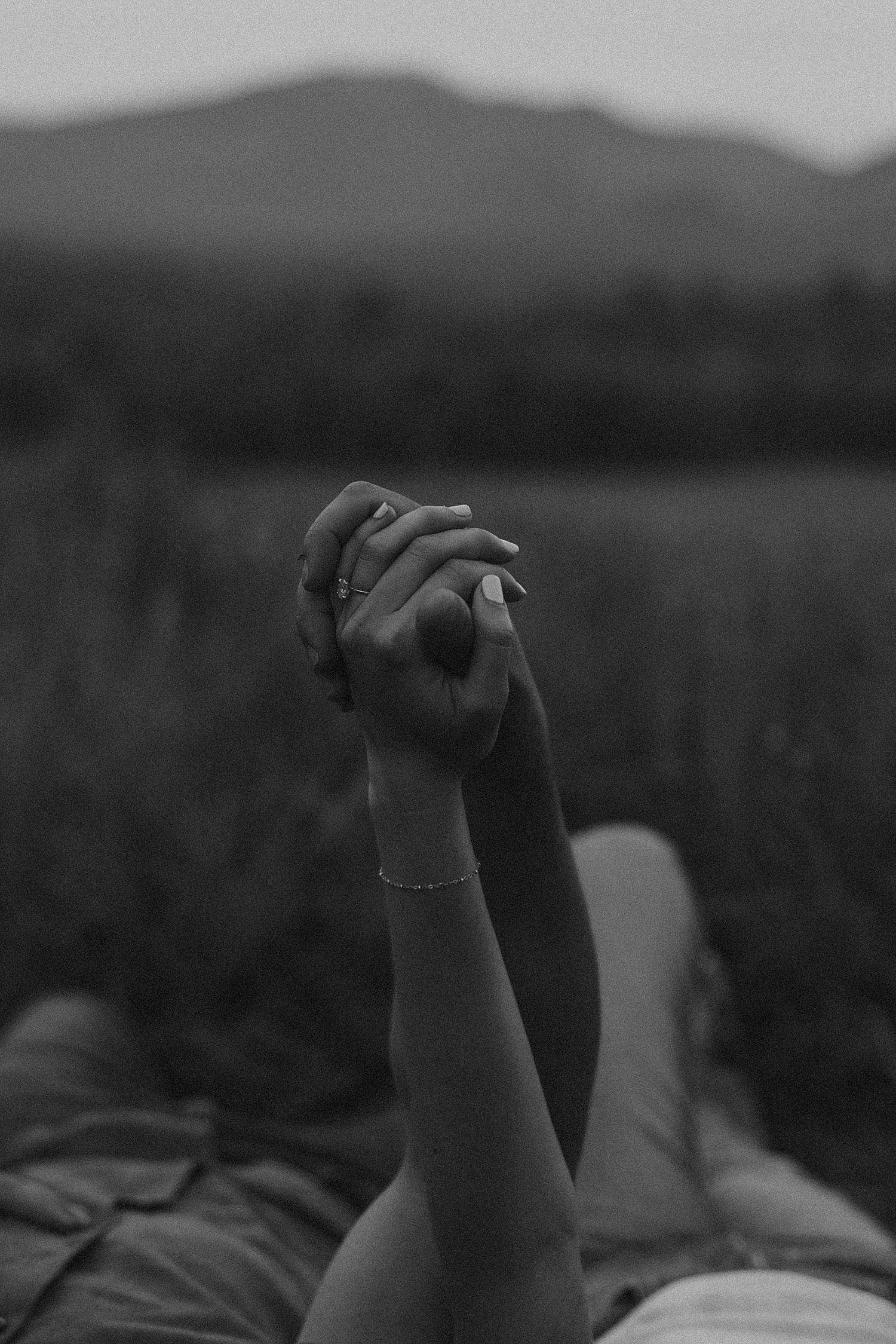 Black and white photo of two people holding hands raised up, with a bracelet visible, set against a blurred field backdrop.