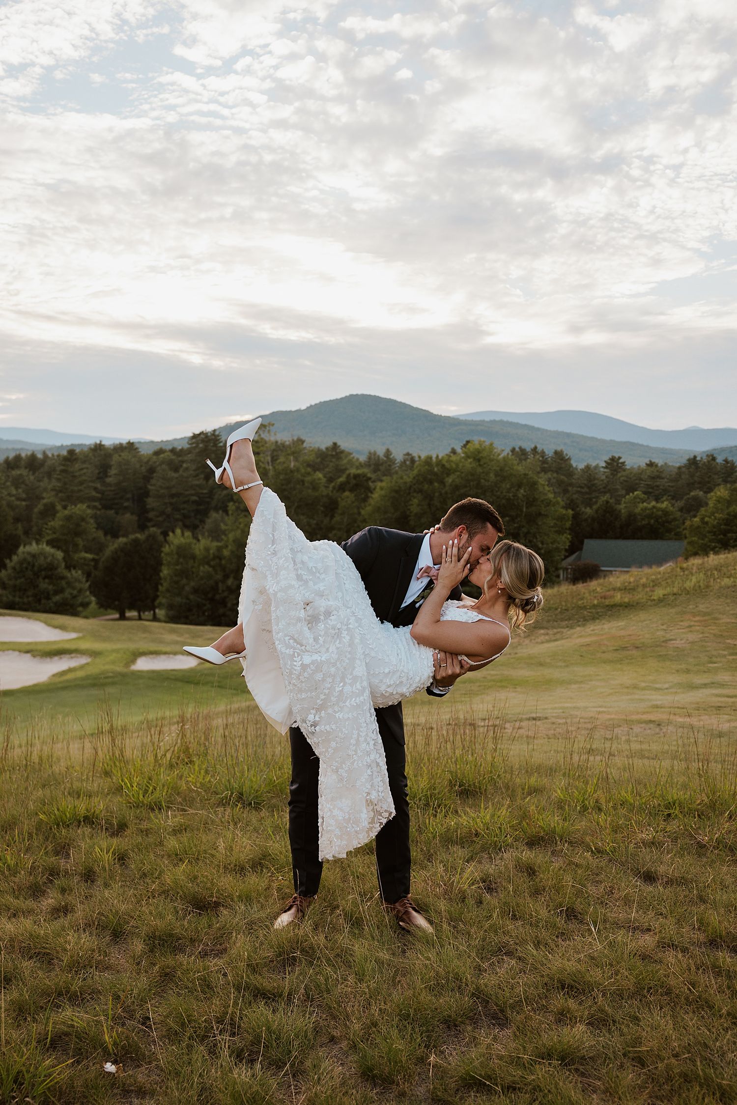 Groom lifts bride in white lace gown for a romantic kiss on a green hillside with views of the White Mountains at the Owl's Nest Resort