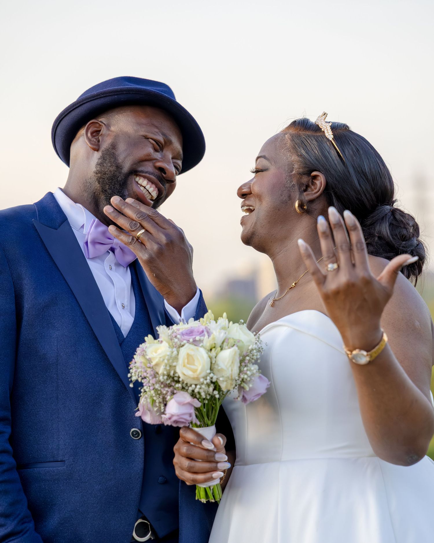 Joyful newlywed couple laughing together, groom in blue suit with bow tie, bride in white gown holding floral bouquet.