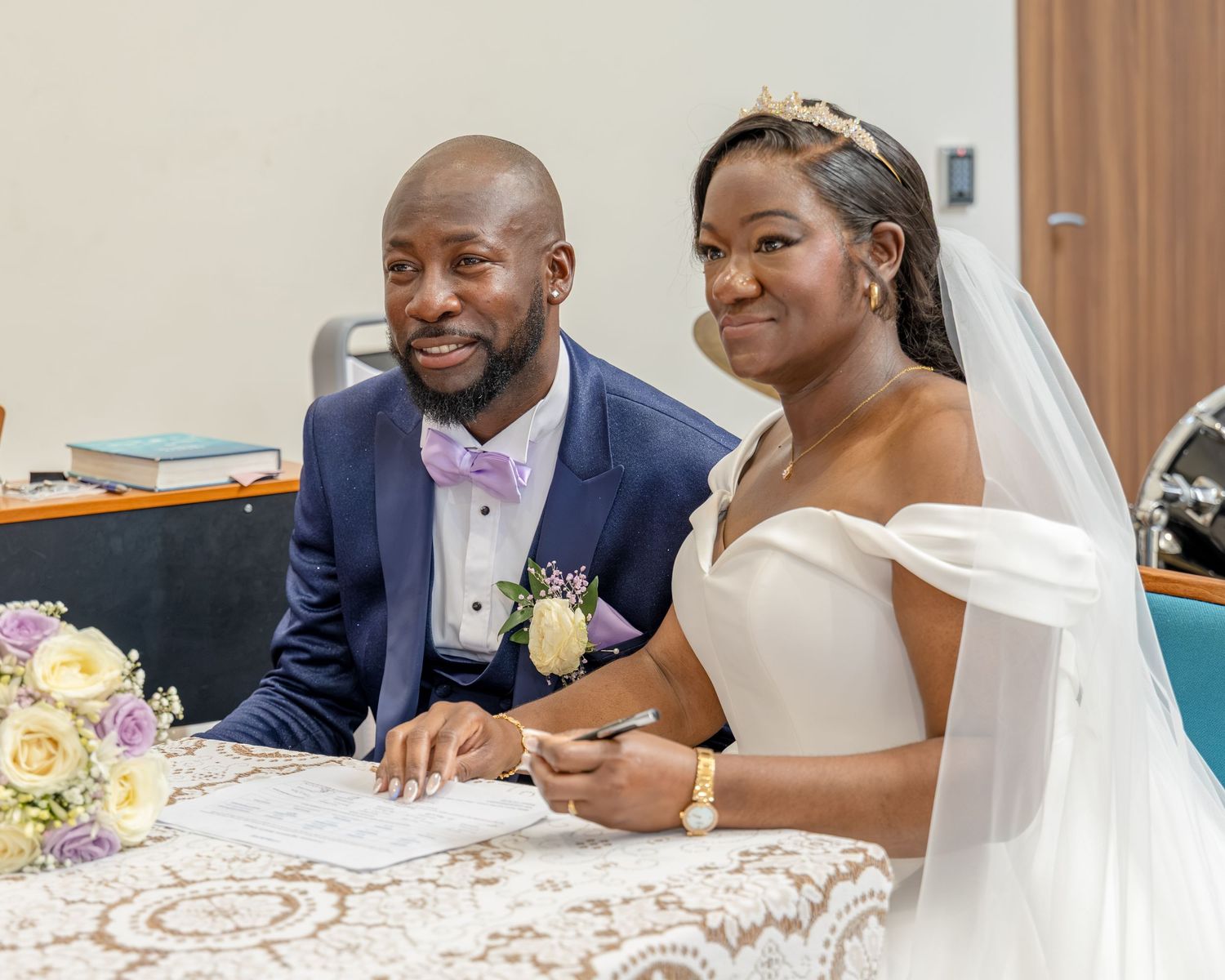 Smiling newlyweds sign wedding documents, groom in blue suit with pink bow tie, bride in white off-shoulder gown and veil.