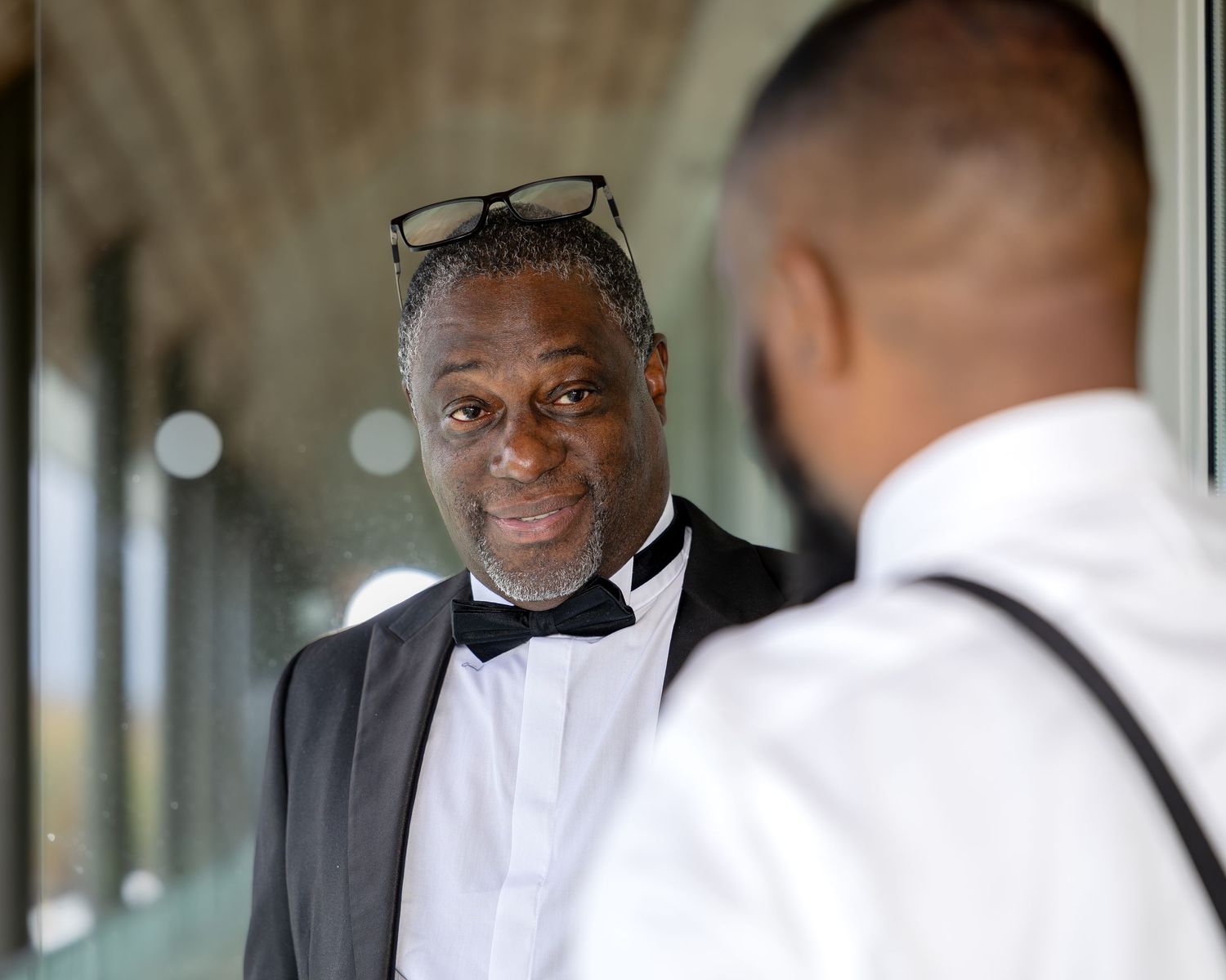 Older gentleman in black and white tuxedo with glasses on head smiling at another person outdoors.