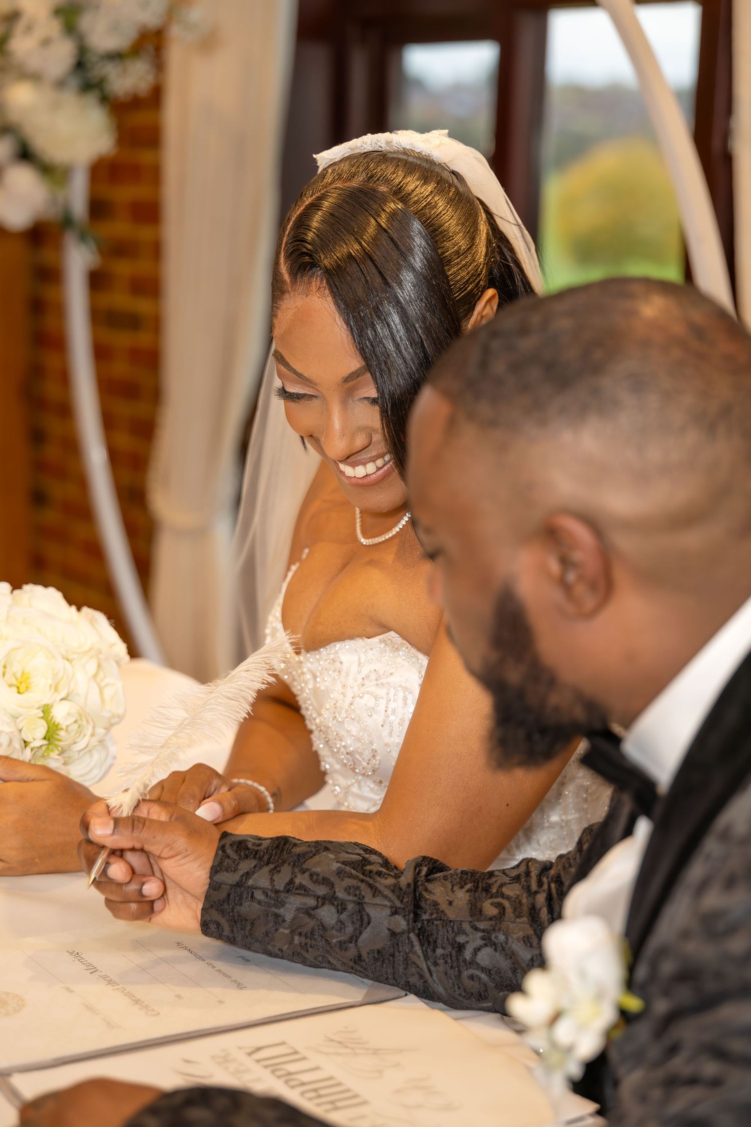 A radiant bride in a lace strapless gown smiles while signing the marriage certificate alongside the groom at a table.