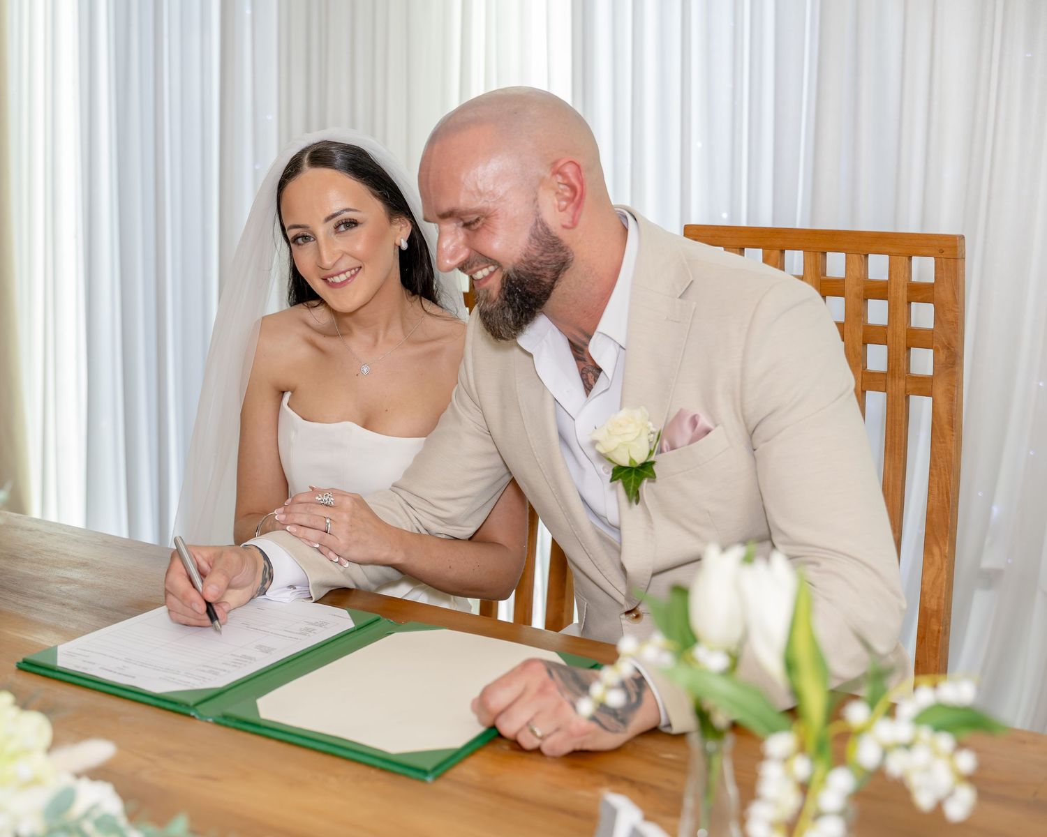 Happy couple signs wedding register at wooden table with white flowers in foreground.