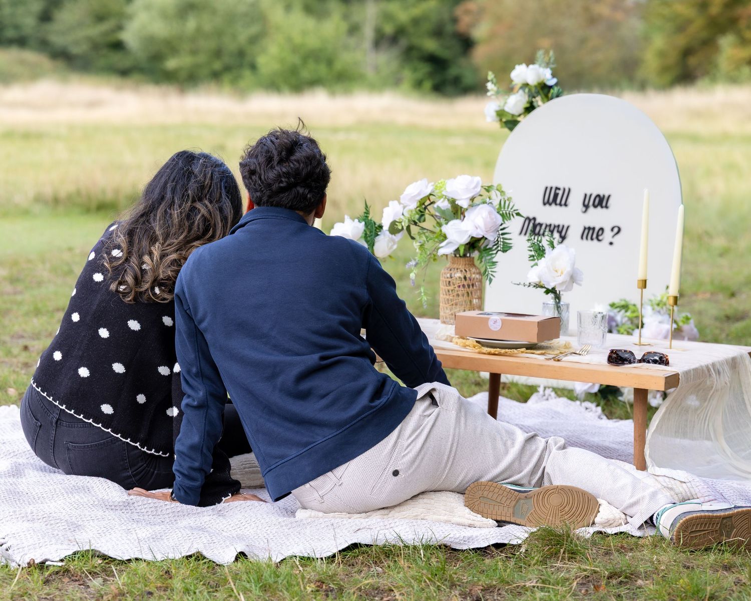 Couple sitting together viewing Will You Marry Me sign at romantic outdoor picnic proposal setup.