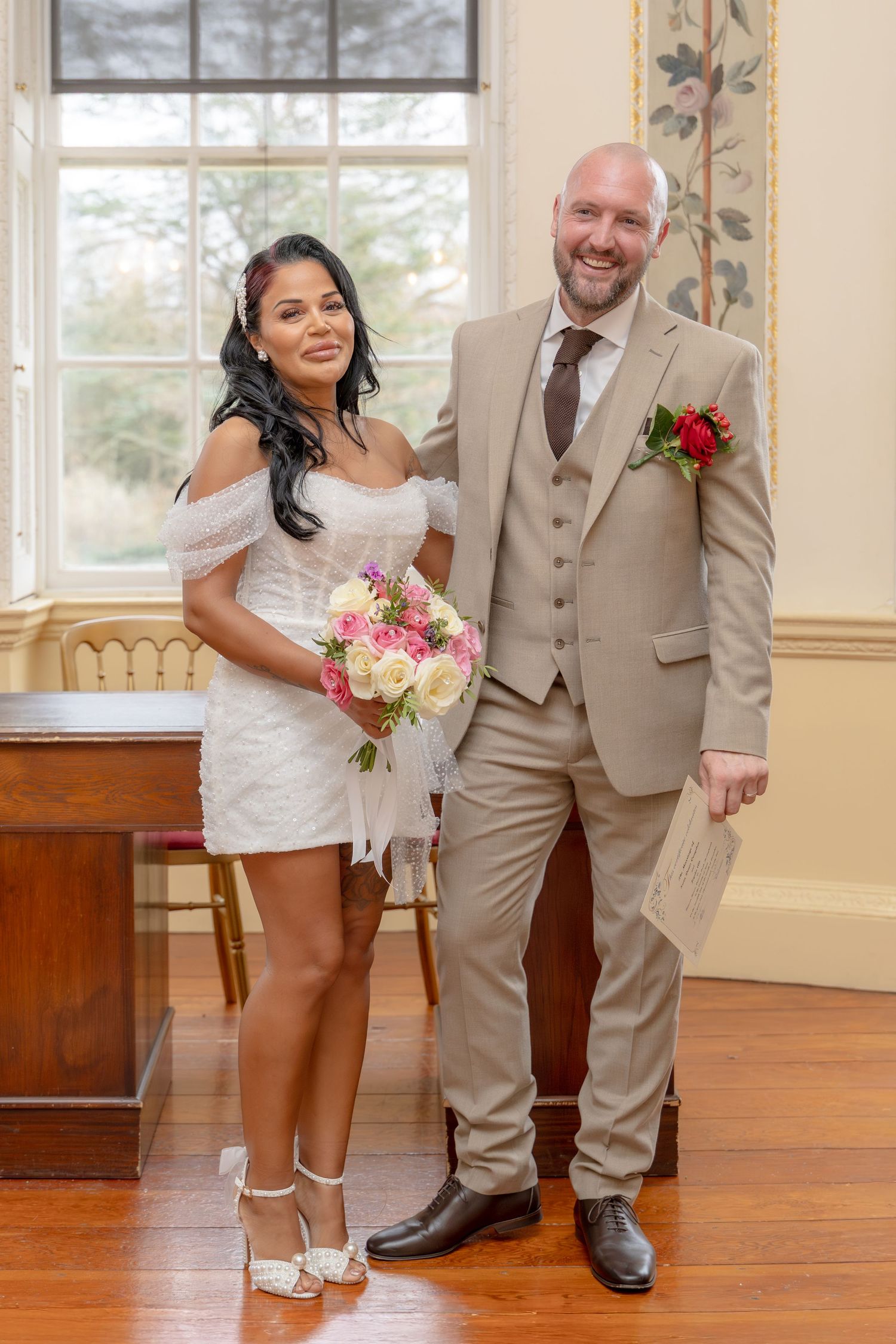 Newlywed couple pose indoors, bride holding pink and white bouquet in short white dress.