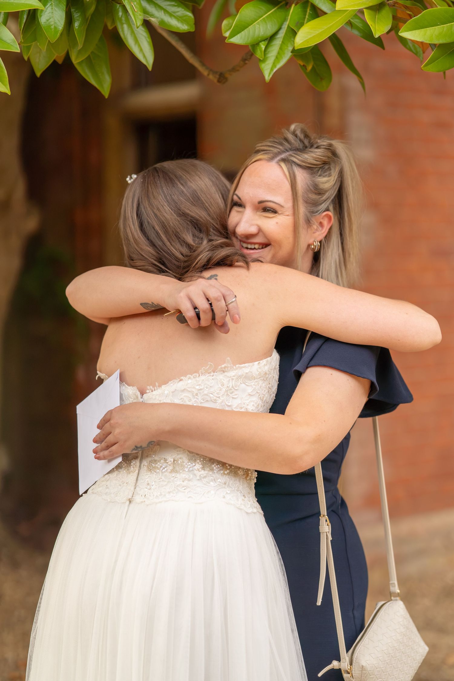 A bride in a white lace gown shares a warm hug with a smiling guest in a navy dress at a wedding.