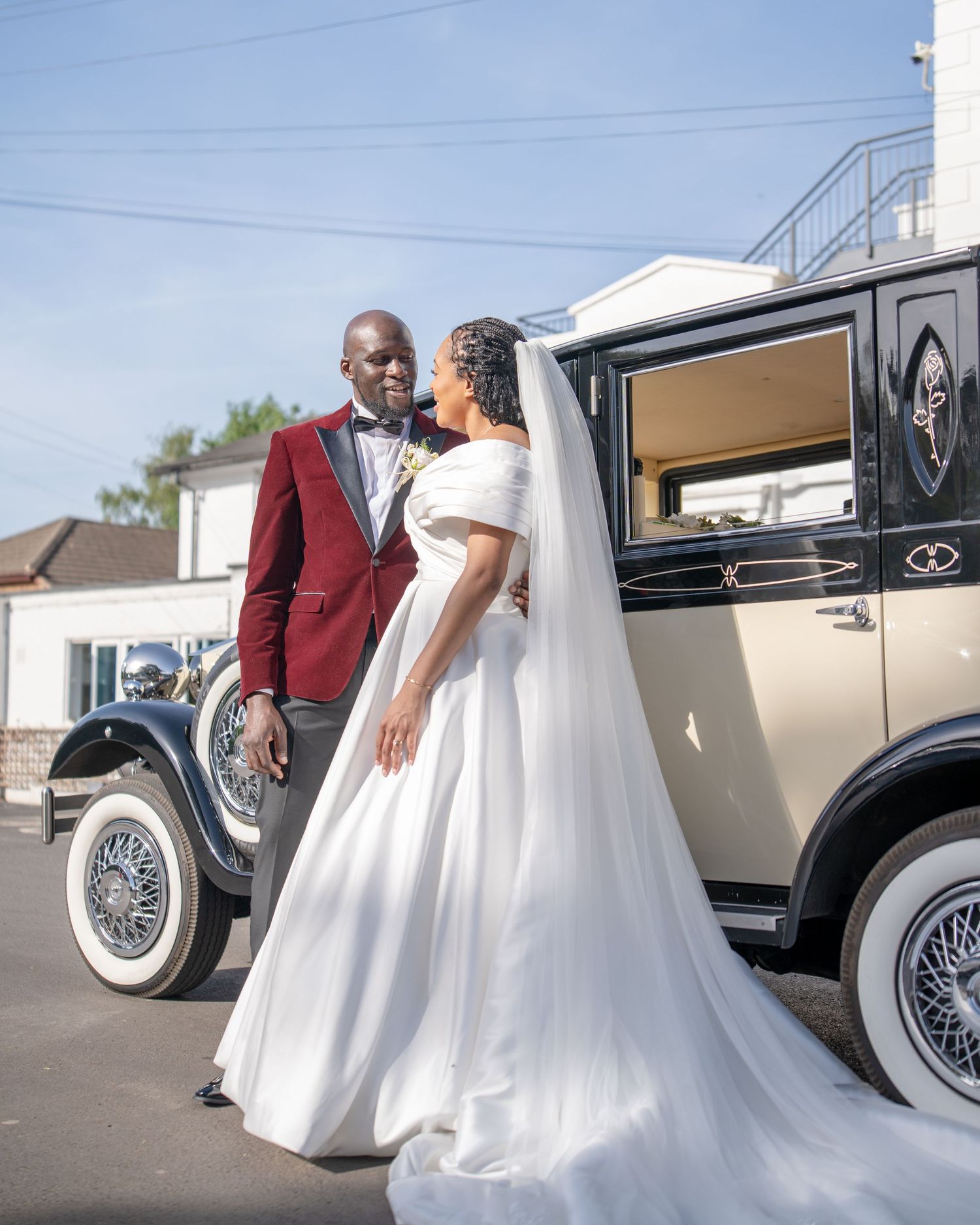Elegant bride and groom posing beside a vintage classic car on their wedding day, groom in burgundy tuxedo.