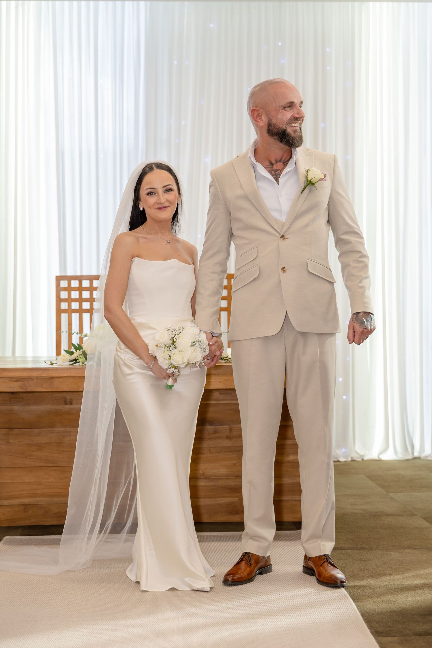 Bride in strapless satin gown and veiled hair stands with tattooed bald groom at wedding altar.