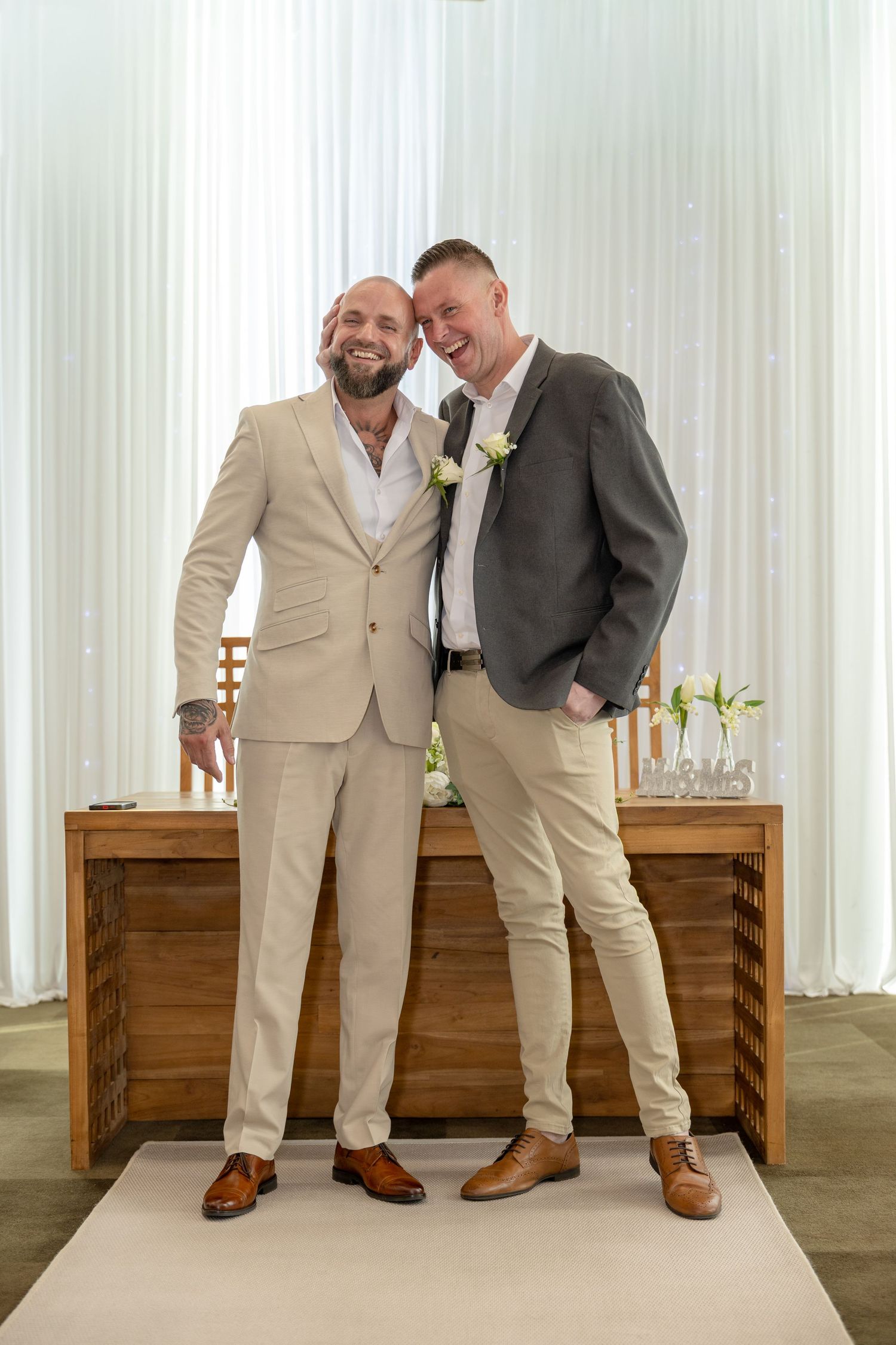 Two smiling men in suits posing together at a wedding ceremony altar.