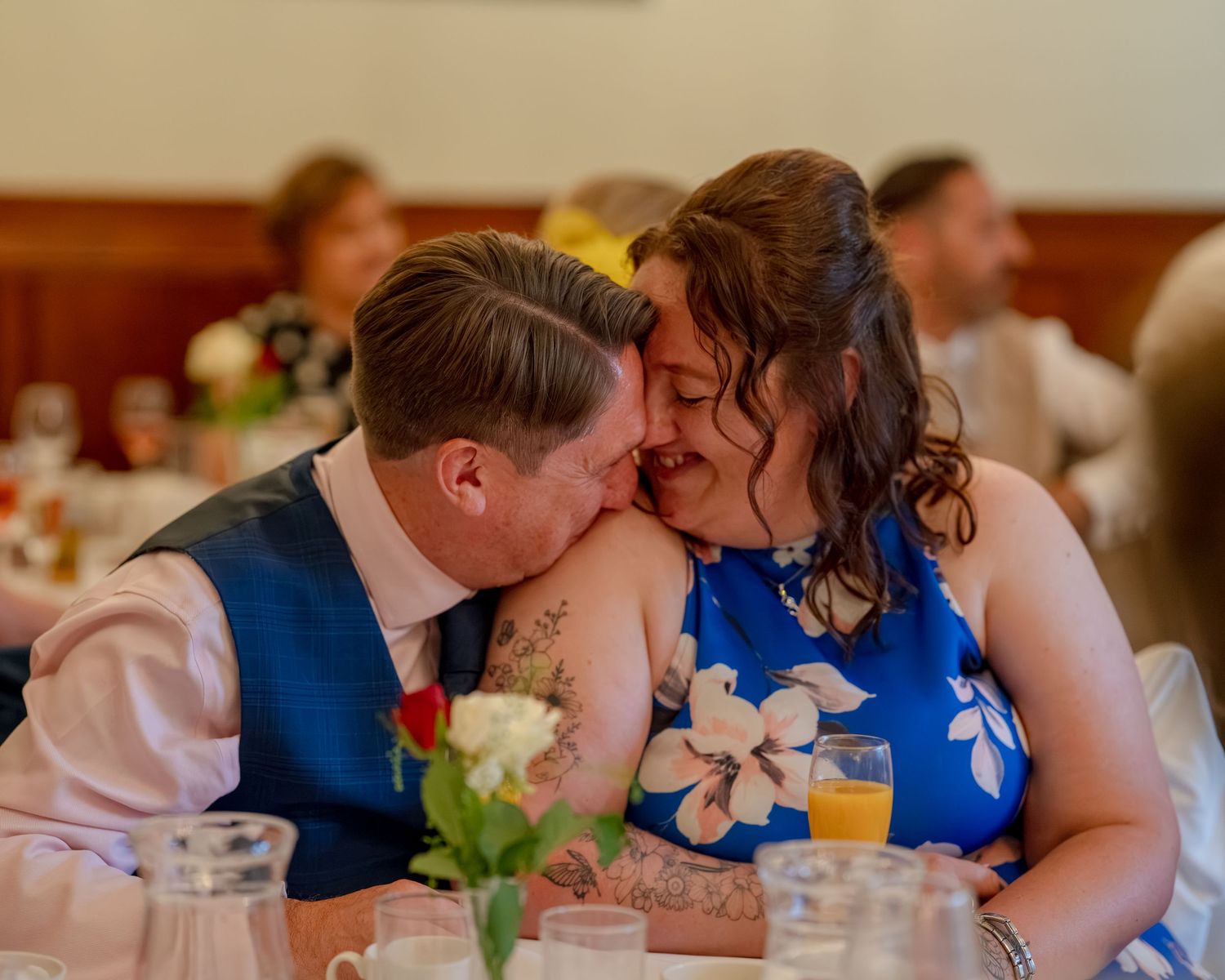 Couple sharing a romantic moment at a wedding reception, woman in blue floral dress, man in blue vest.