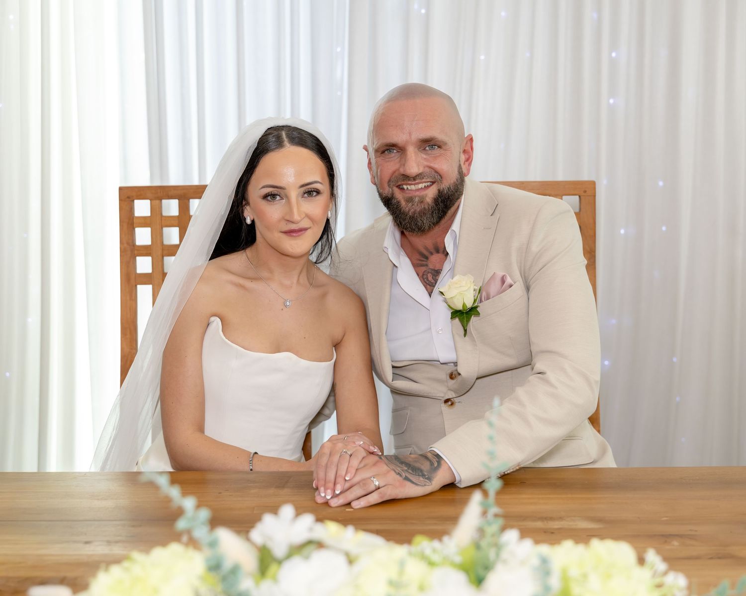 Bride in veil and groom in beige suit pose together at wedding signing table.