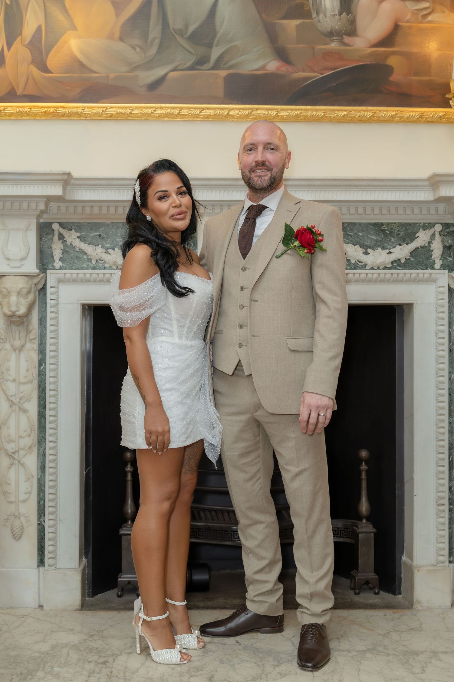 Bride and groom posing together in front of ornate fireplace, bride in short white dress, groom in tan suit.