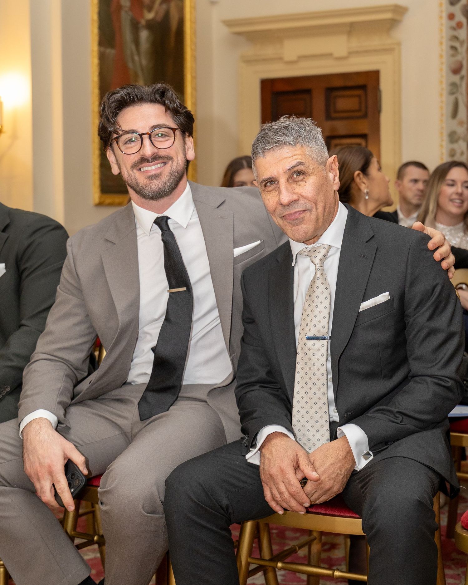 Two smiling men in grey suits seated together at a formal wedding reception.