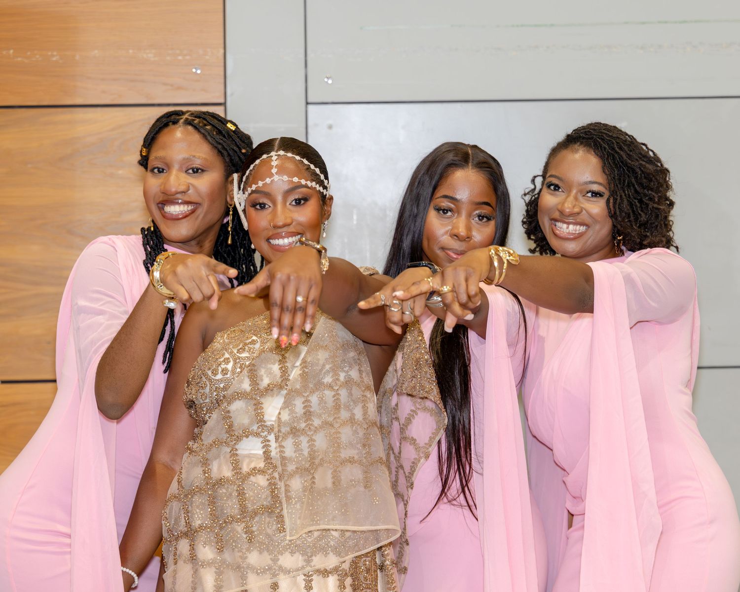 Bride in gold sequin dress poses with three bridesmaids in pink gowns pointing at wedding rings.