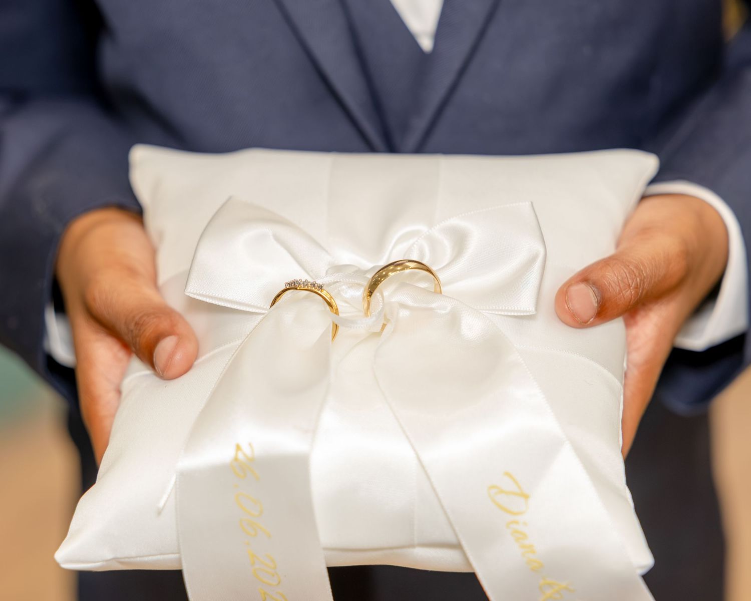 Gold wedding rings resting on a white satin ring bearer pillow with bow, held by a man in a blue suit.