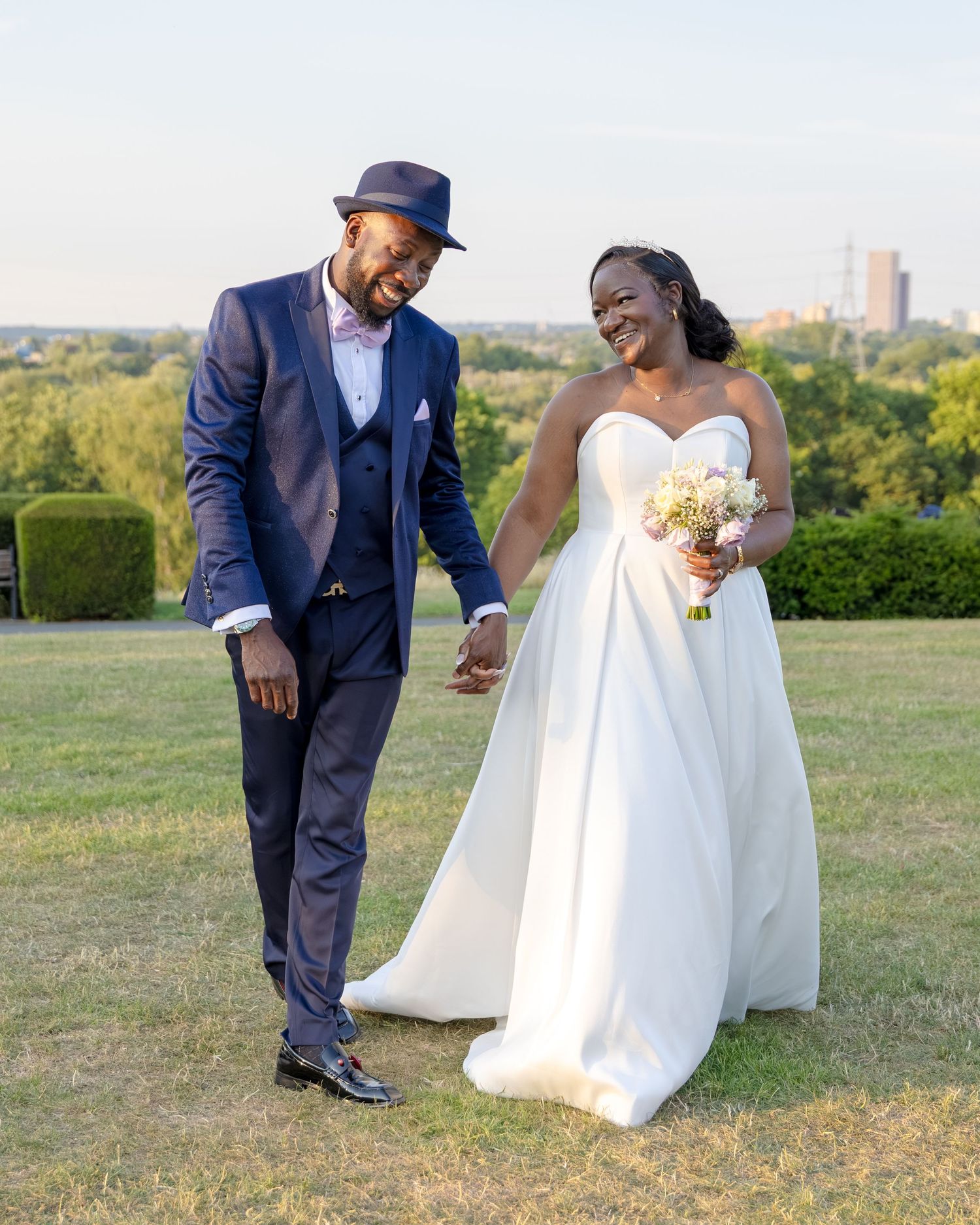 A joyful Black couple in wedding attire walks hand-in-hand on a green lawn, groom in navy suit, bride in white gown.