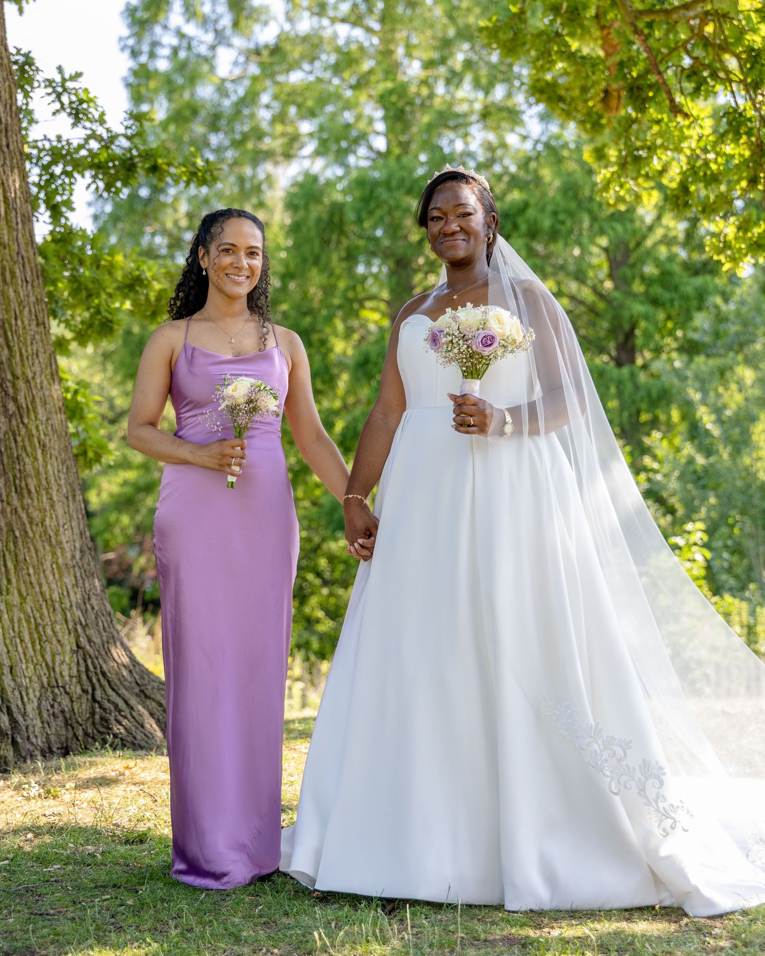 Bride in white gown and bridesmaid in purple dress holding bouquets outdoors among green trees.