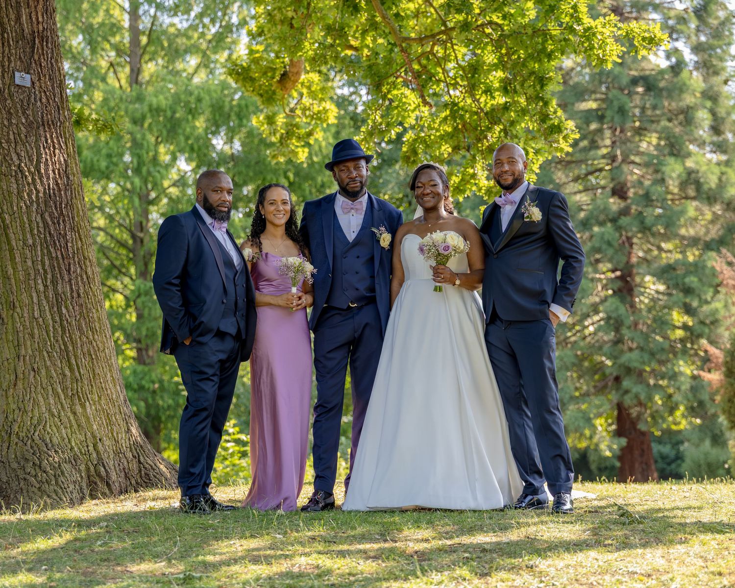 Wedding party poses outdoors under green trees, bride in white gown with bouquet, groom in blue suit, groomsmen and bridesmaid.