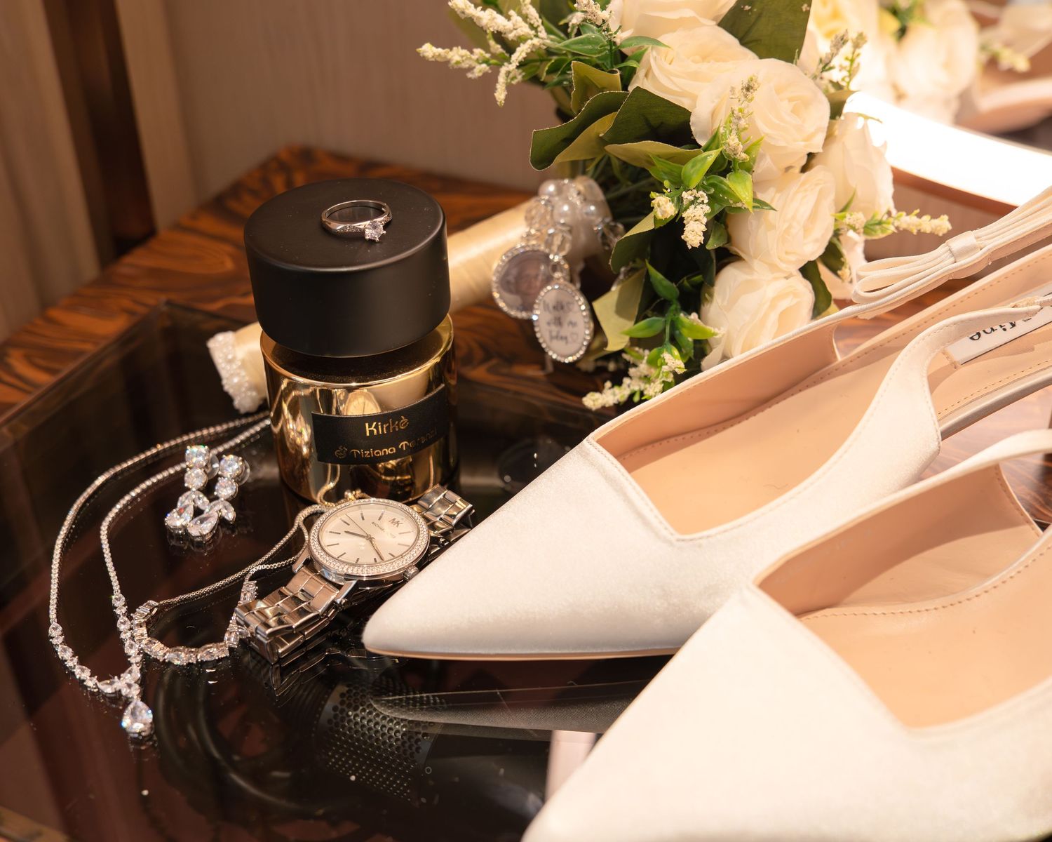 Bridal flat shoes, perfume, watch, diamond necklace, earrings, and white rose bouquet arranged on a wooden surface.