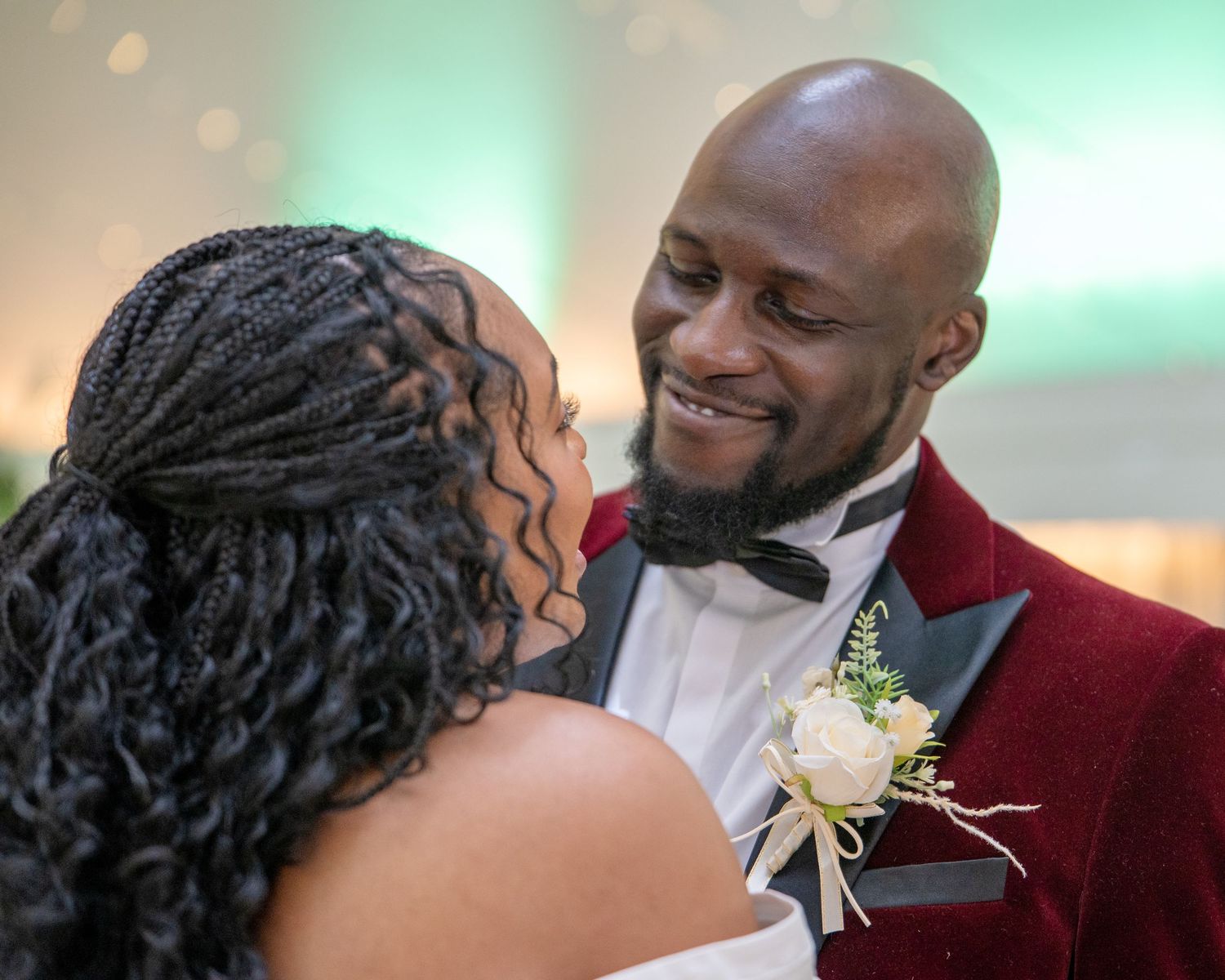 A bride and groom share a joyful moment, groom wearing a red velvet tuxedo with boutonniere at their wedding reception.
