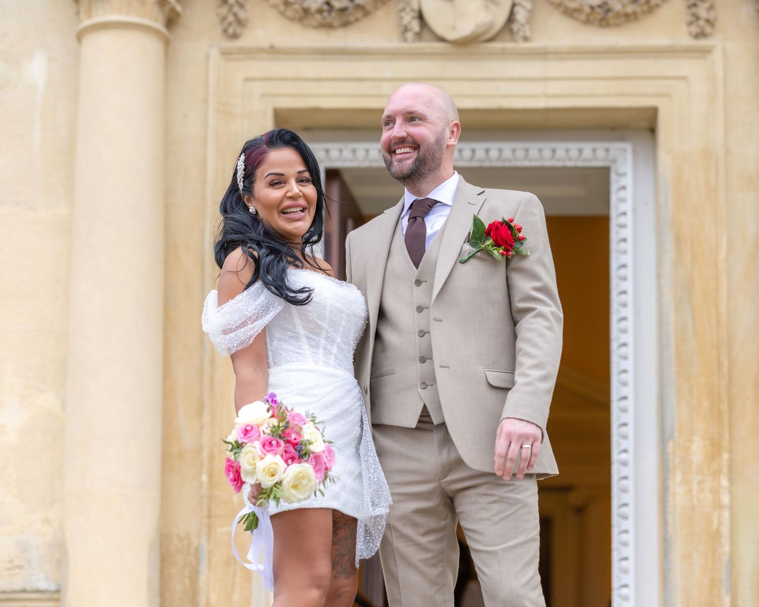 Bride and groom smile outdoors at stone building, bride holding colorful bouquet in white dress.