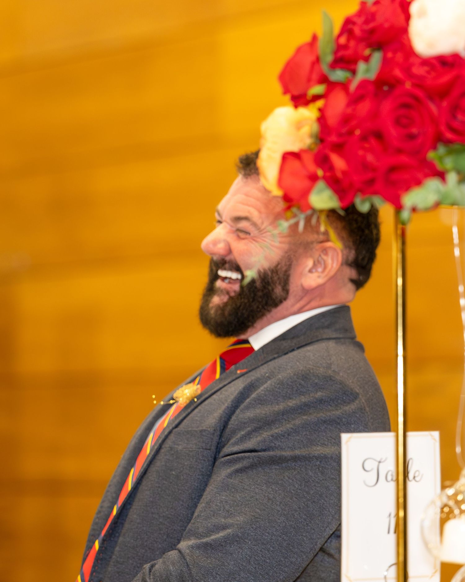 Bearded man in gray suit laughing joyfully at a wedding reception near red roses.