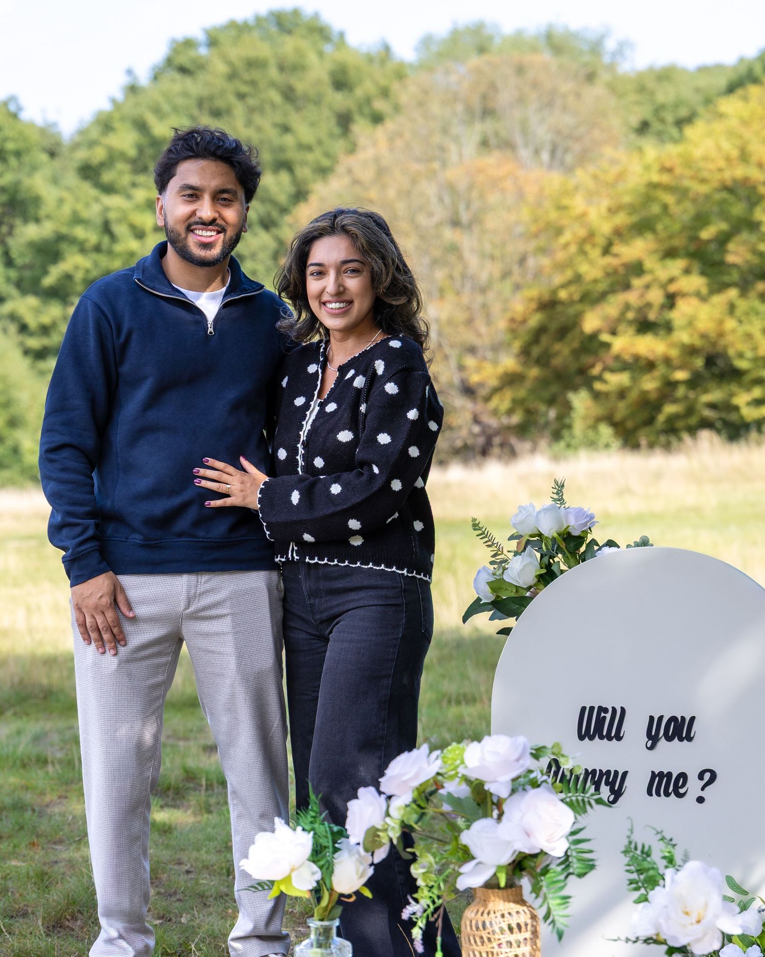 Happy couple standing together next to a Will You Marry Me sign with white flowers outdoors.