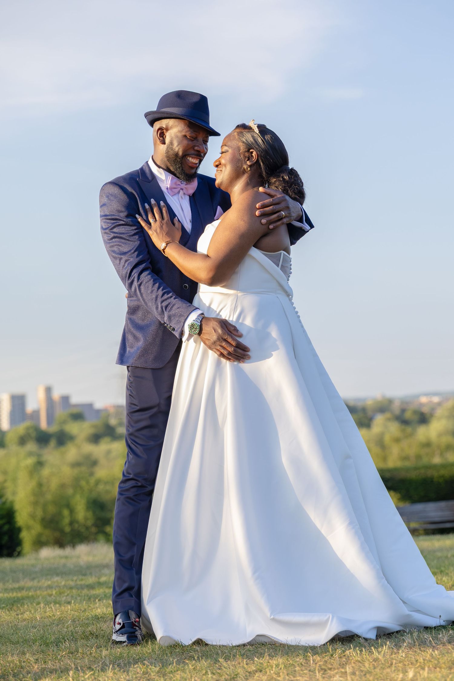 A joyful Black couple embraces outdoors on their wedding day, groom in navy suit and hat, bride in white gown.
