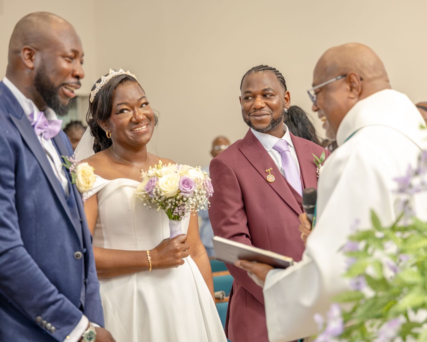 Bride smiling with bouquet at wedding ceremony, flanked by groom and guests, as officiant reads vows.