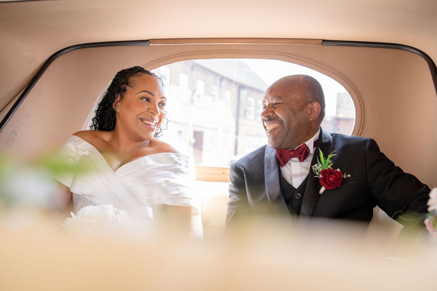 Bride and groom smiling at each other inside a vintage car on their wedding day.